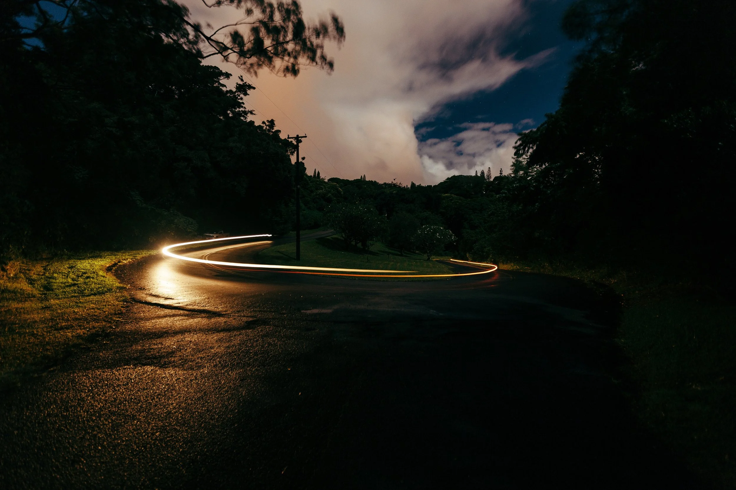 White-and-gold vehicle light trail curving around a wet road beneath a clouded evening sky