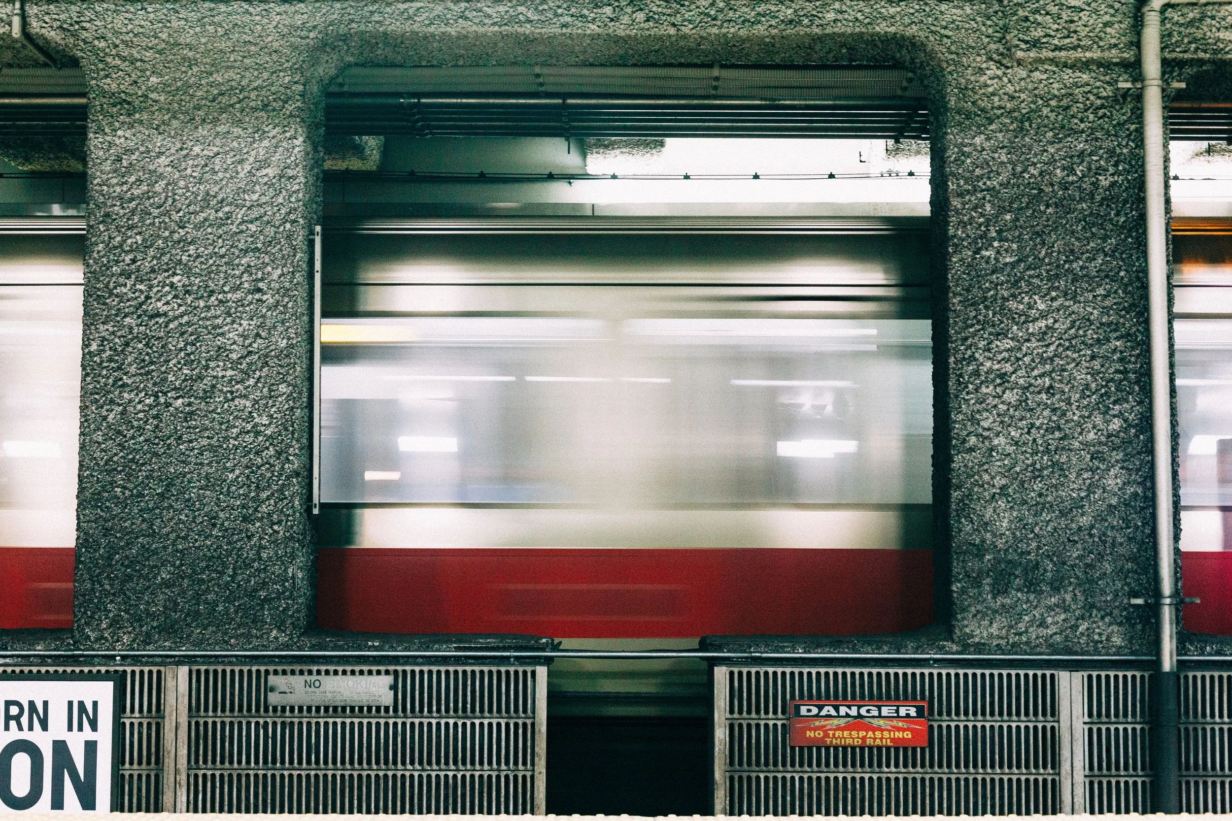 Red-and-white train blurred in motion behind a concrete platform opening and metal railing