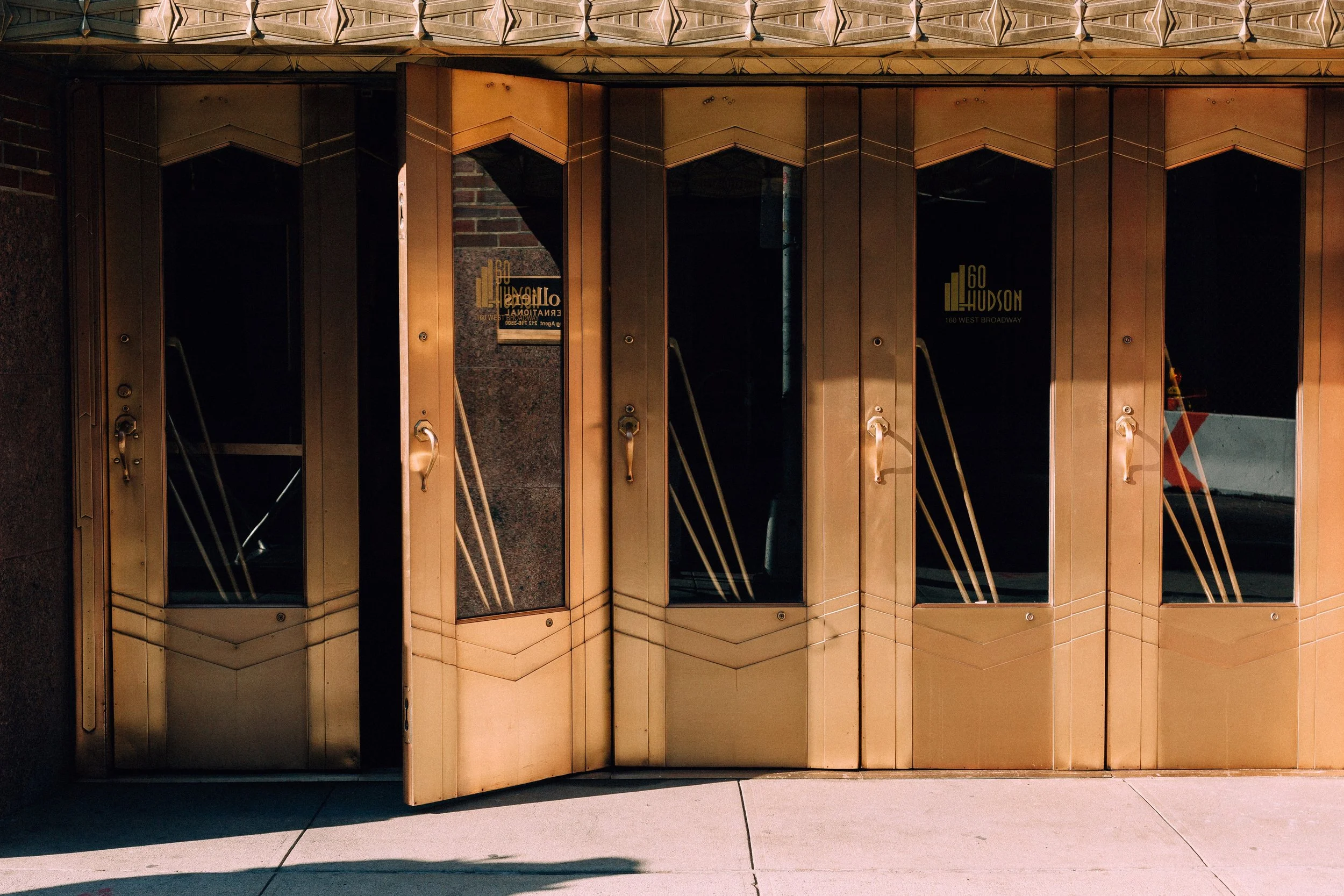 Row of bronze-toned Art Deco doors with one door open into a dark interior in hard sunlight