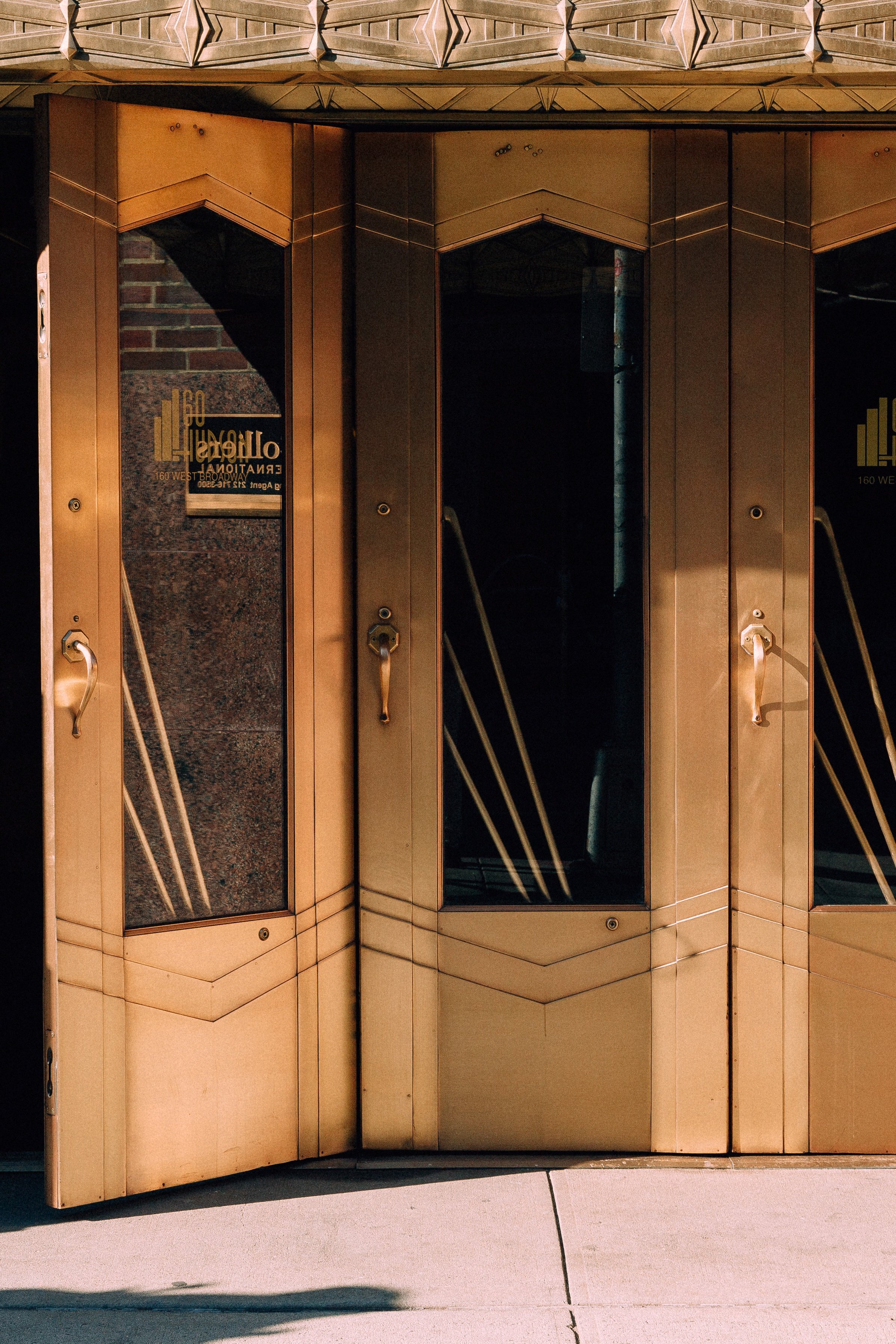 Vertical detail of the open door, geometric glass panels, bronze handles, and sidewalk shadow