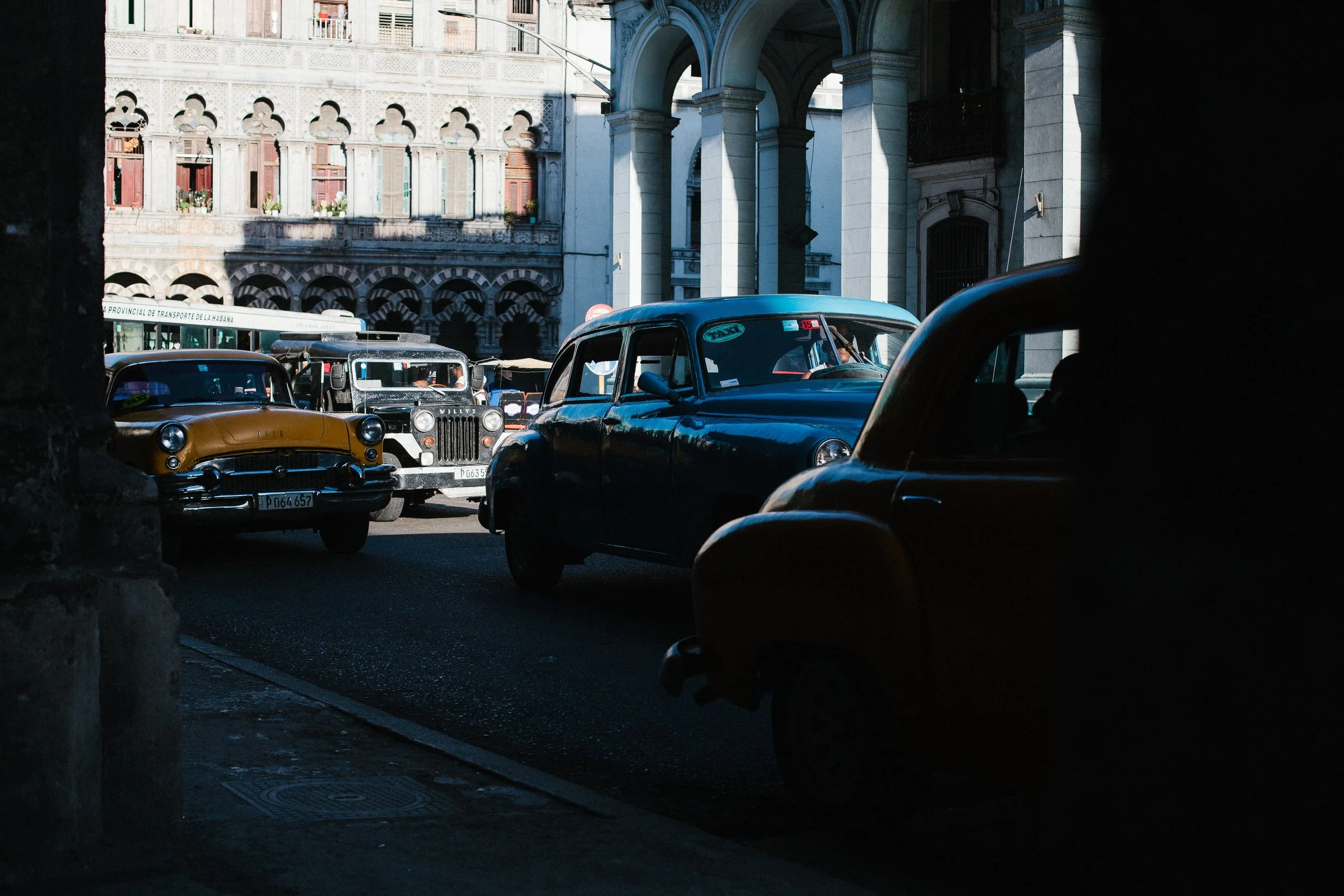 Classic cars moving through deep street shadow beneath arches in Havana, Cuba