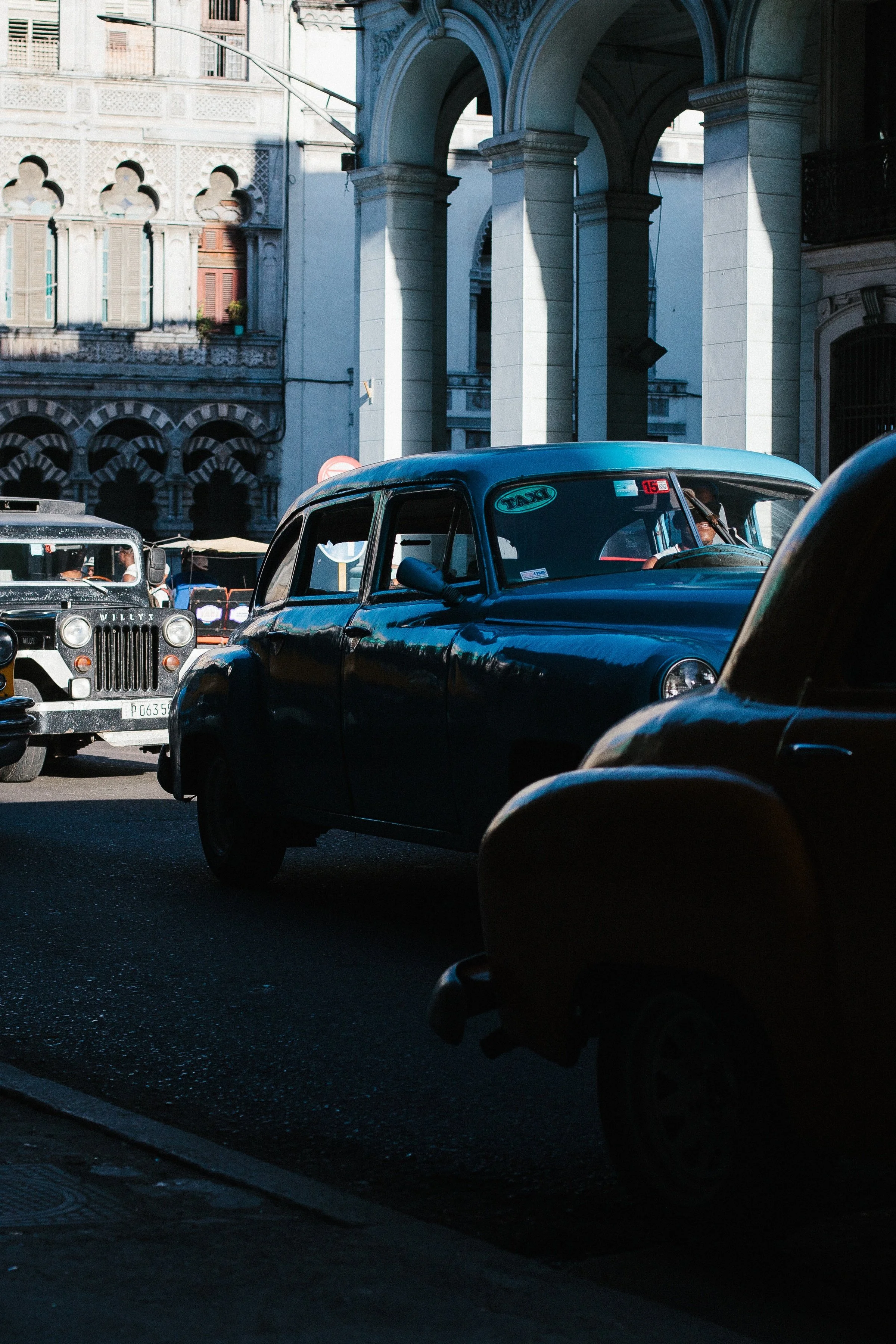 Vertical detail of blue and dark classic cars, chrome, arches, and sunlit Havana architecture