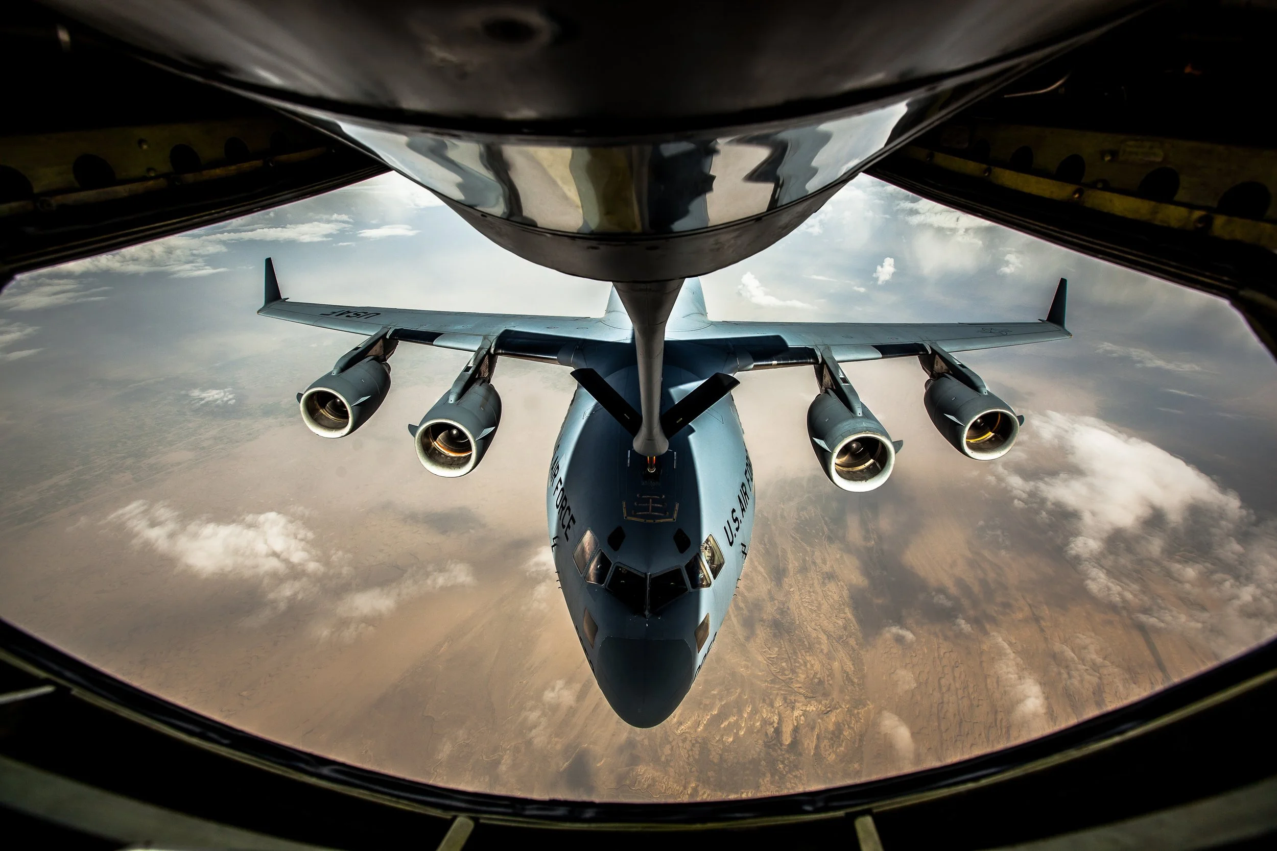 Four-engine military aircraft aligned beneath a refueling boom above clouds and distant ground