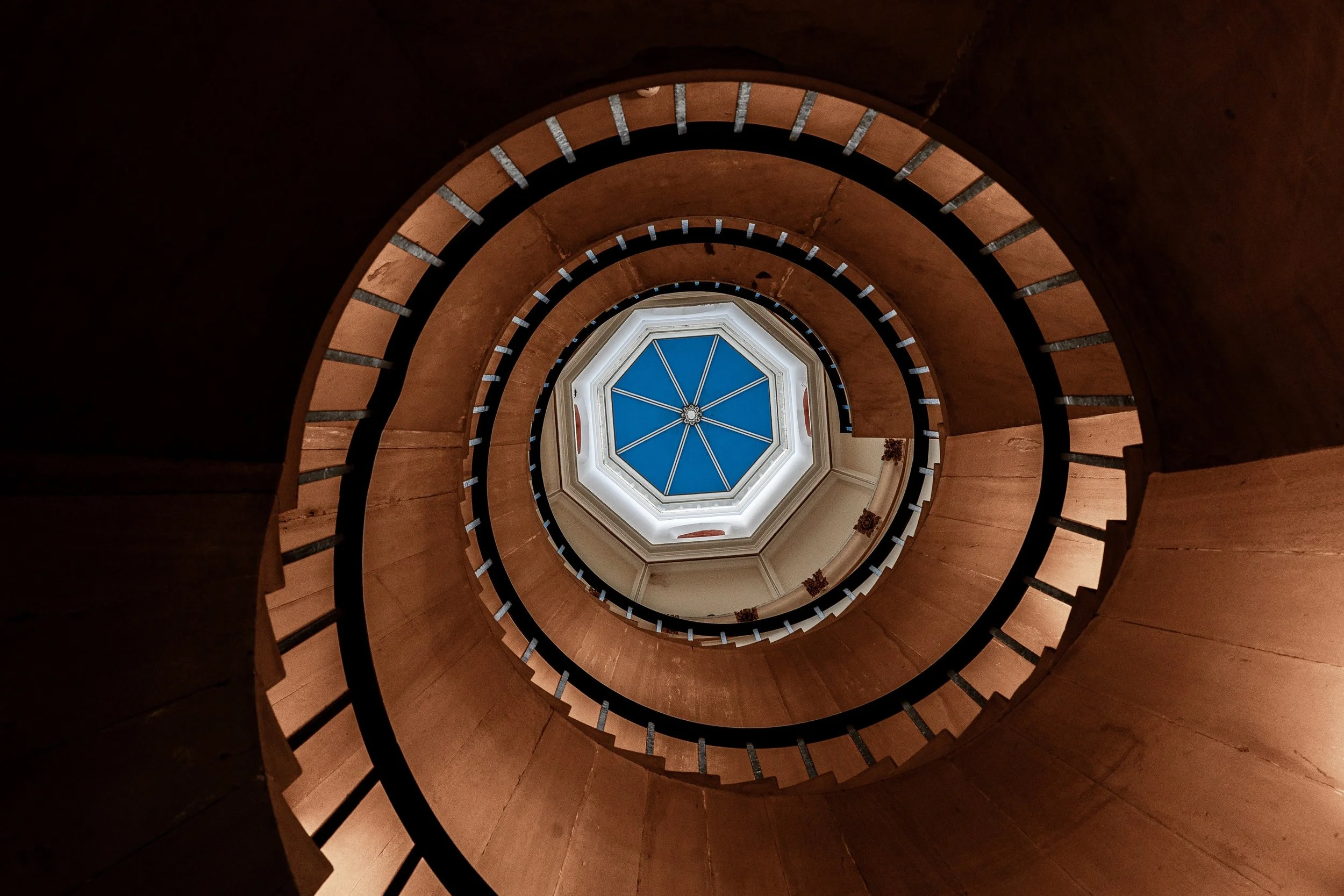 Upward view through warm spiral staircase rings toward a blue octagonal skylight