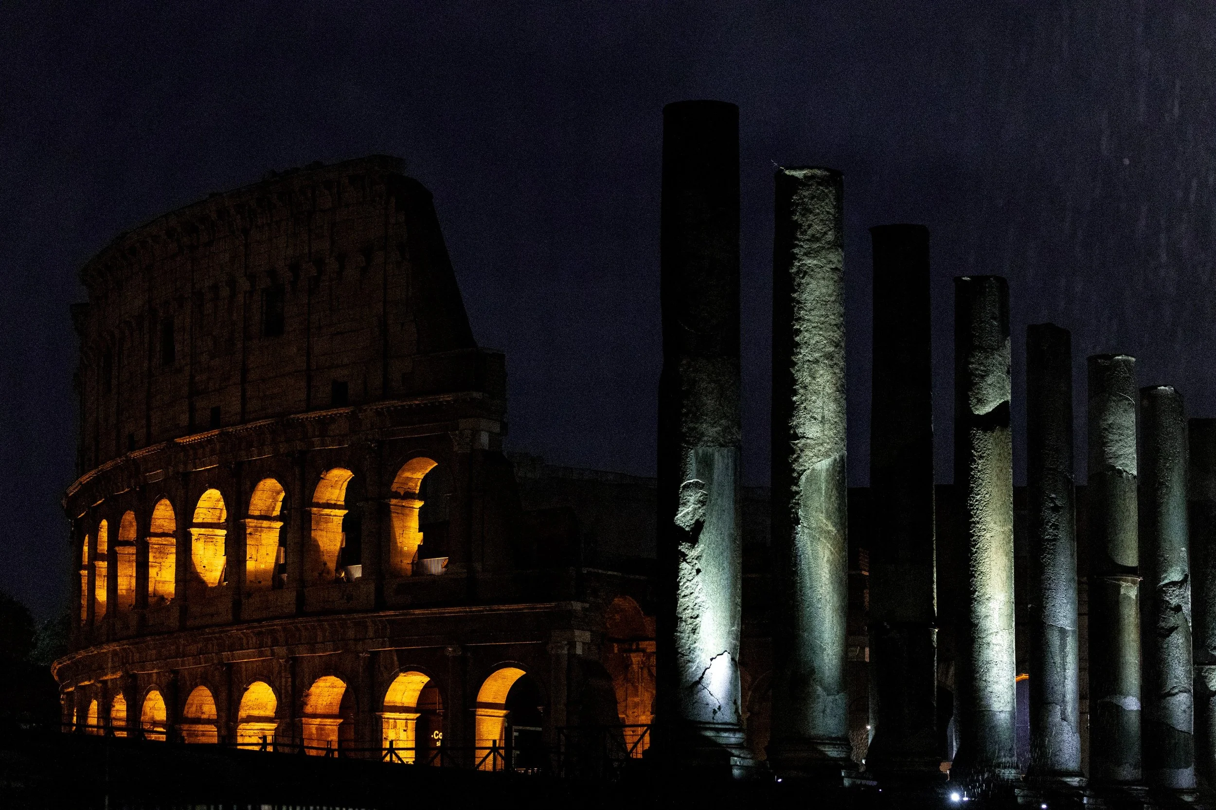 Illuminated Colosseum arches and a row of ancient columns against the dark Roman night