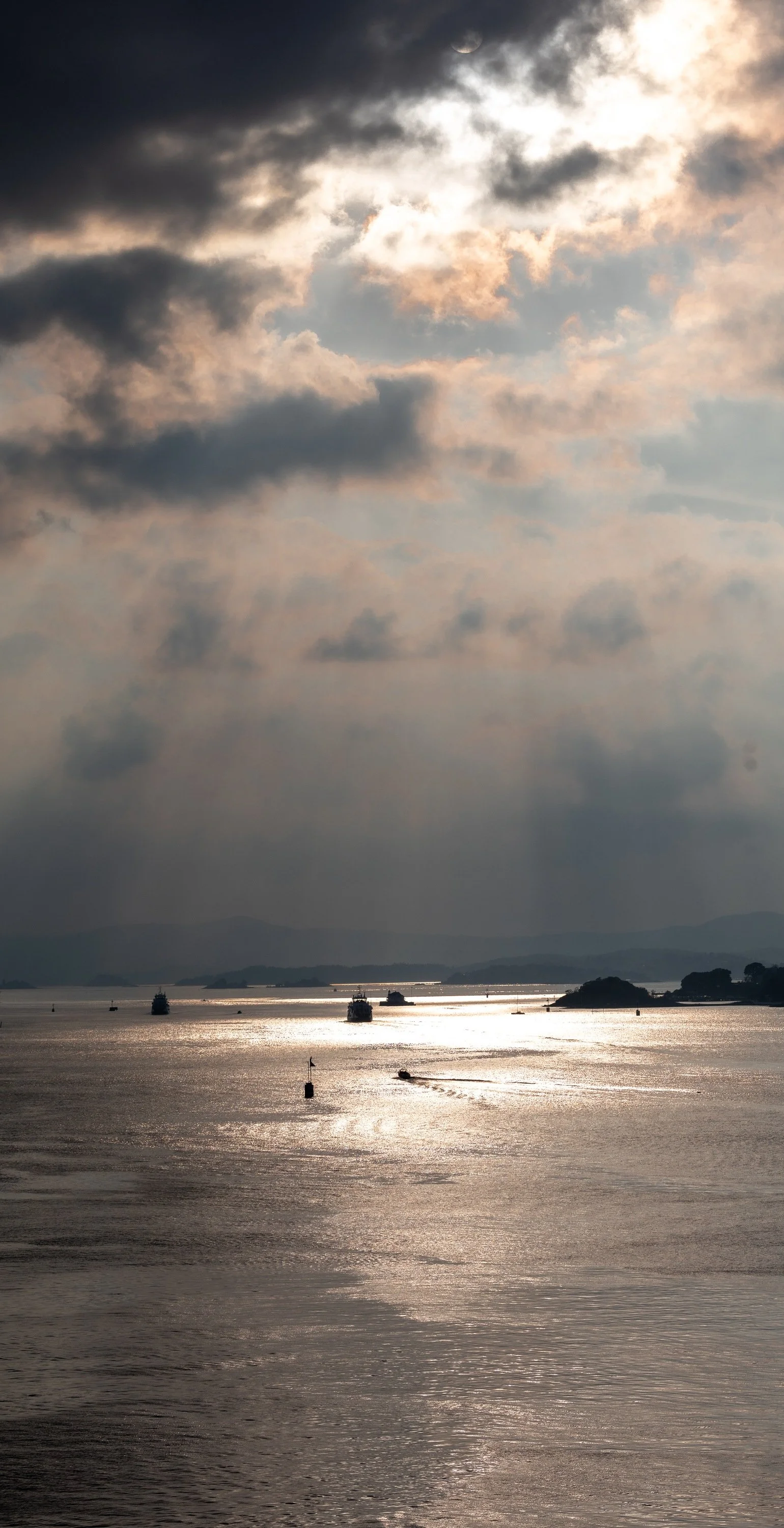 Silver sunlight crossing open water with small boats, rocks, islands, and a hazy coast