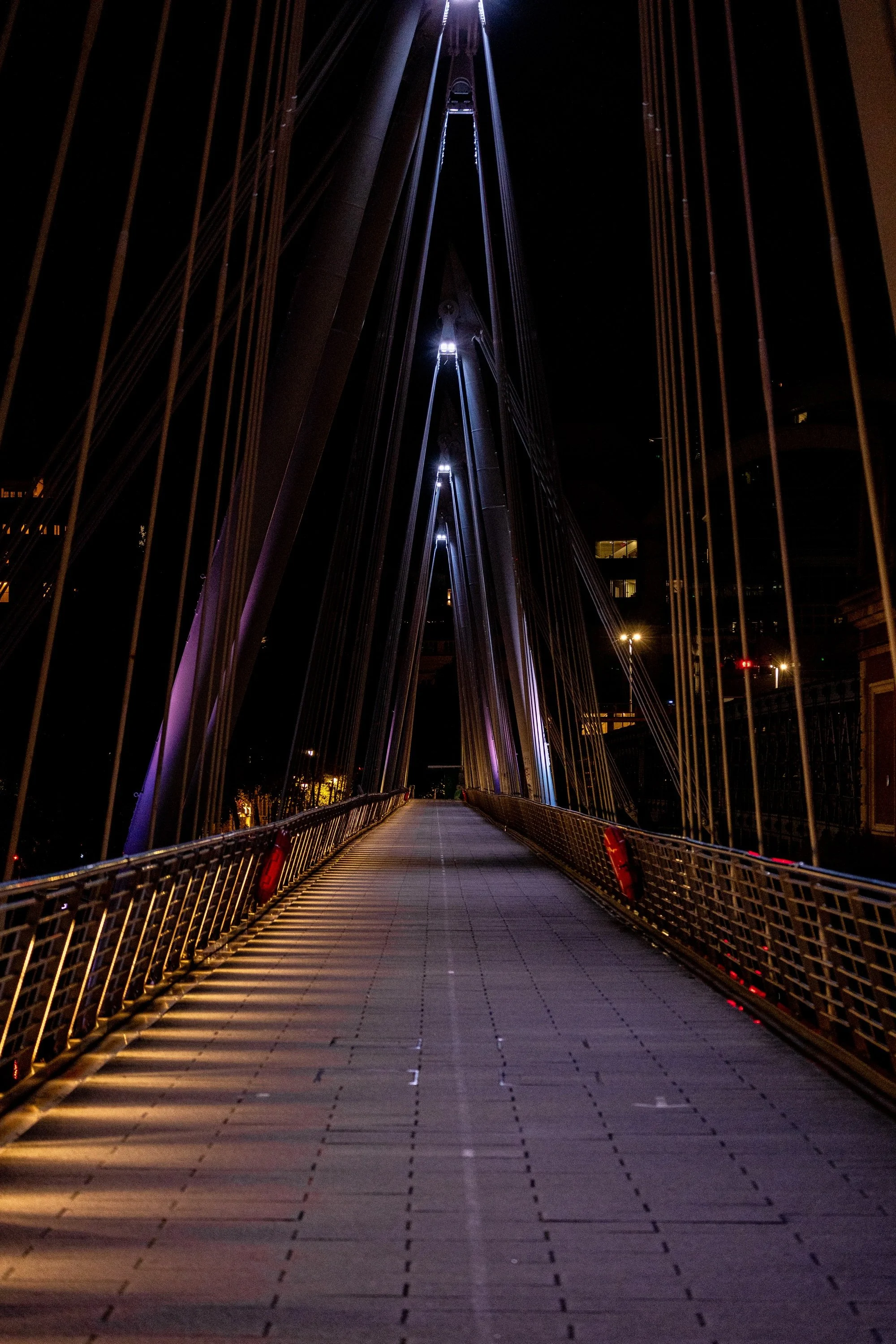 Centered suspension bridge walkway at night beneath repeating towers, cables, and cool blue lights