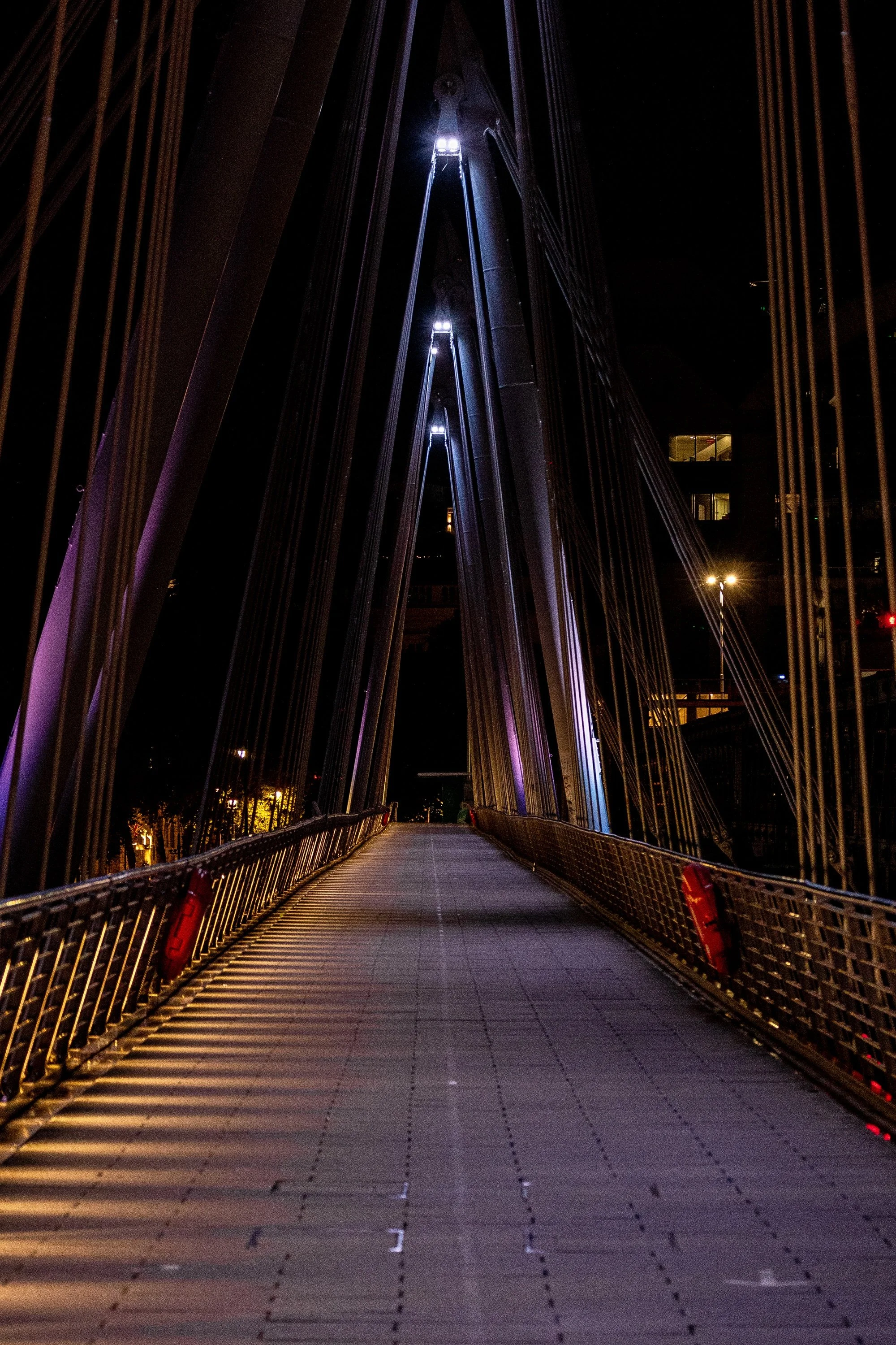 Close detail of illuminated bridge railings, converging cables, and the walkway leading into darkness