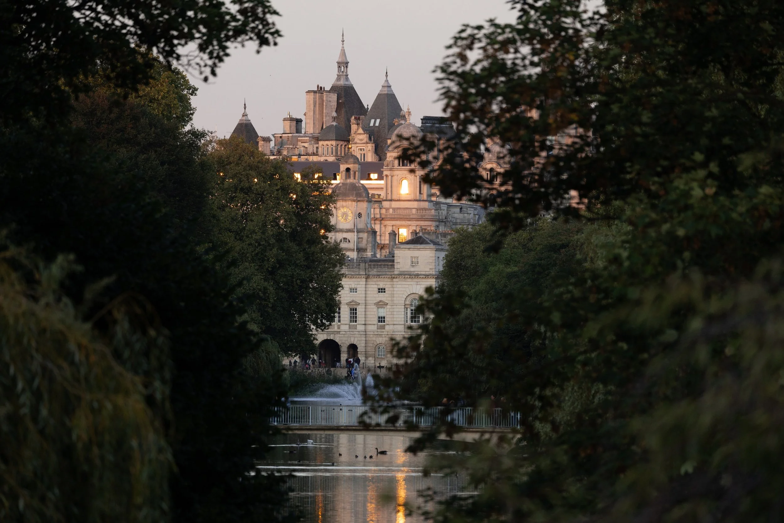 Ornate pale stone London building seen through dense trees across a reflective pond