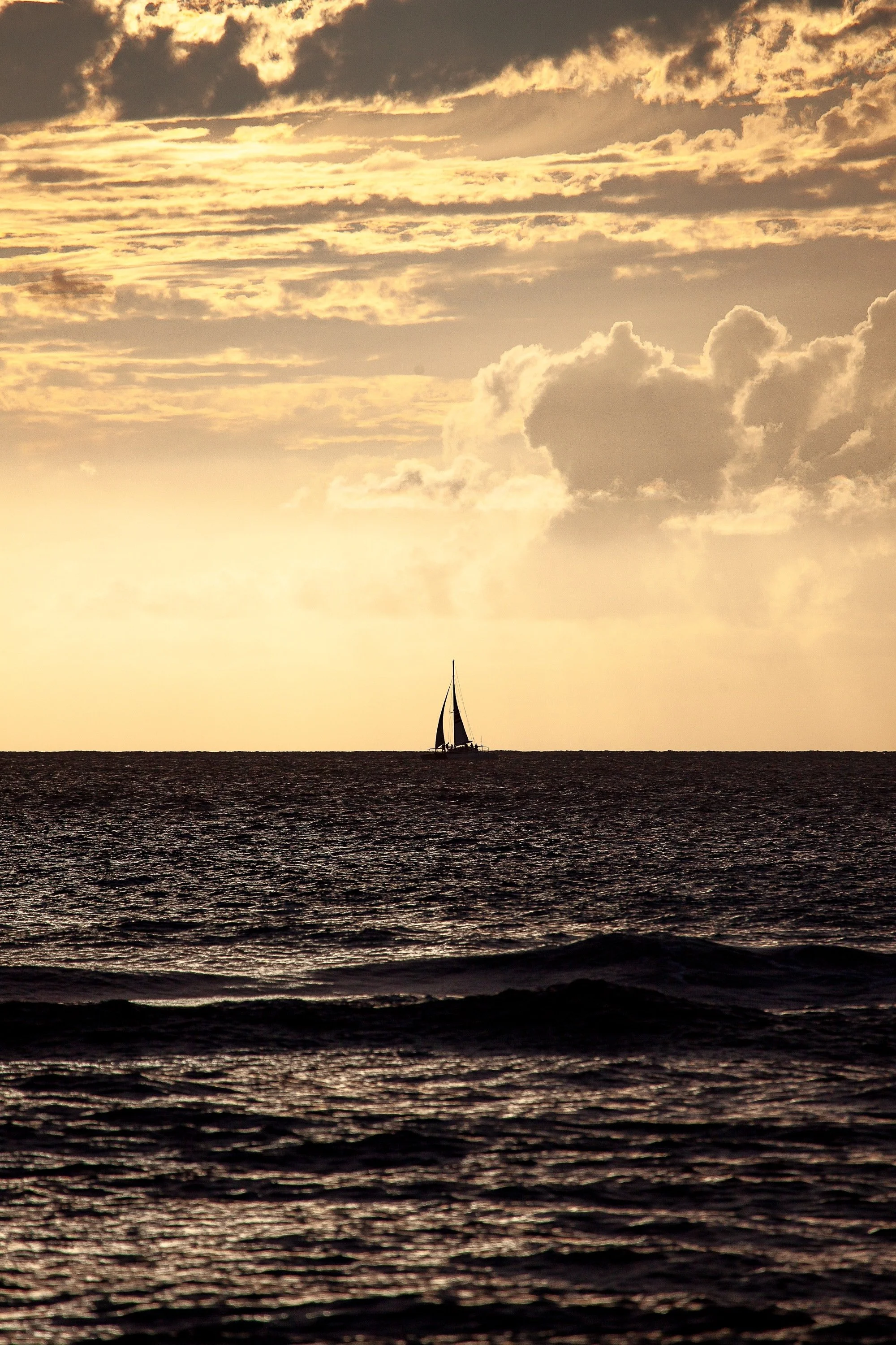 Vertical seascape photograph of a small sailboat on the horizon beneath layered golden clouds on Hawaii's North Shore.