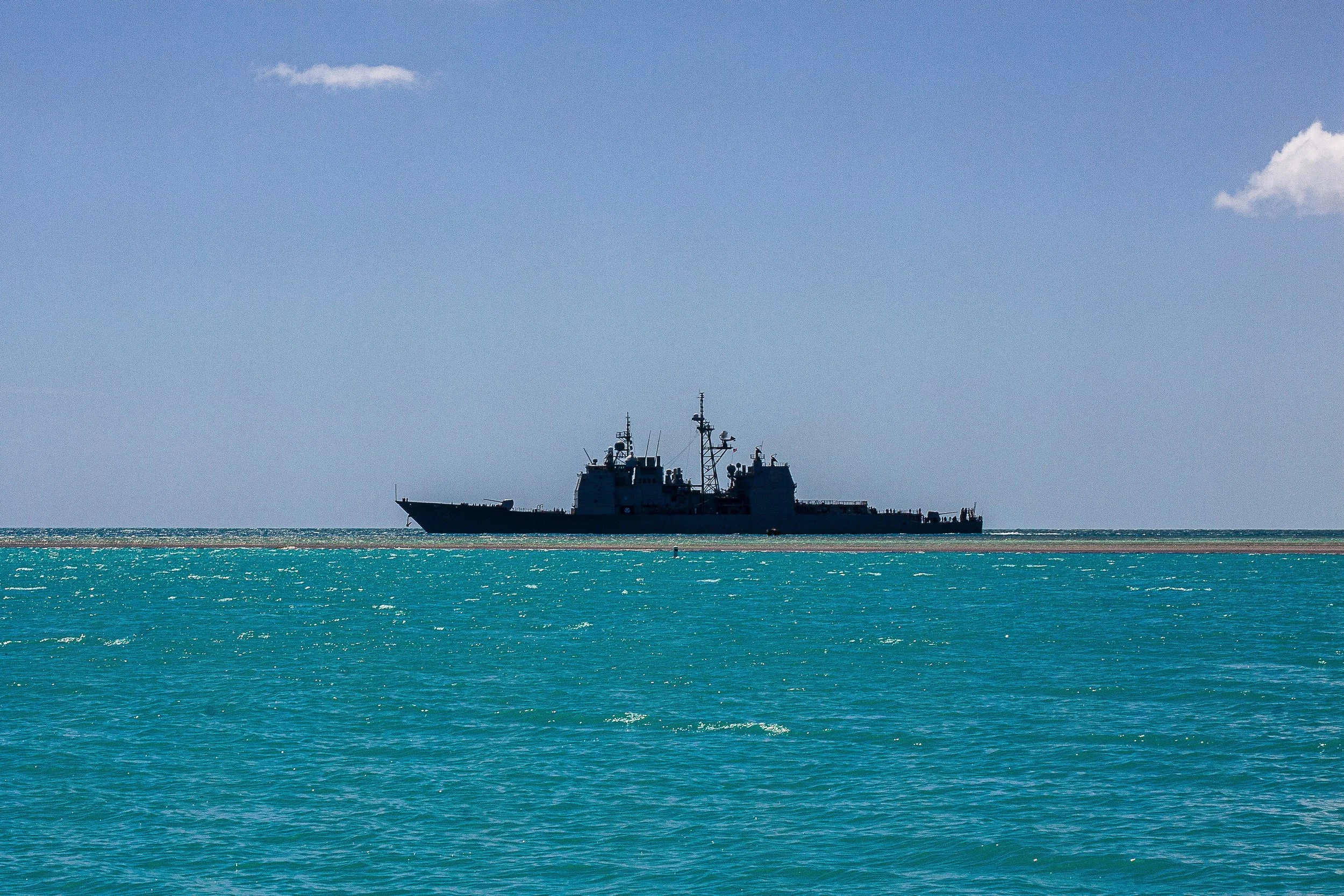 Complete uncropped artwork for Pacific Sentinel, showing a dark naval vessel crosses clear turquoise water beneath a spare blue sky