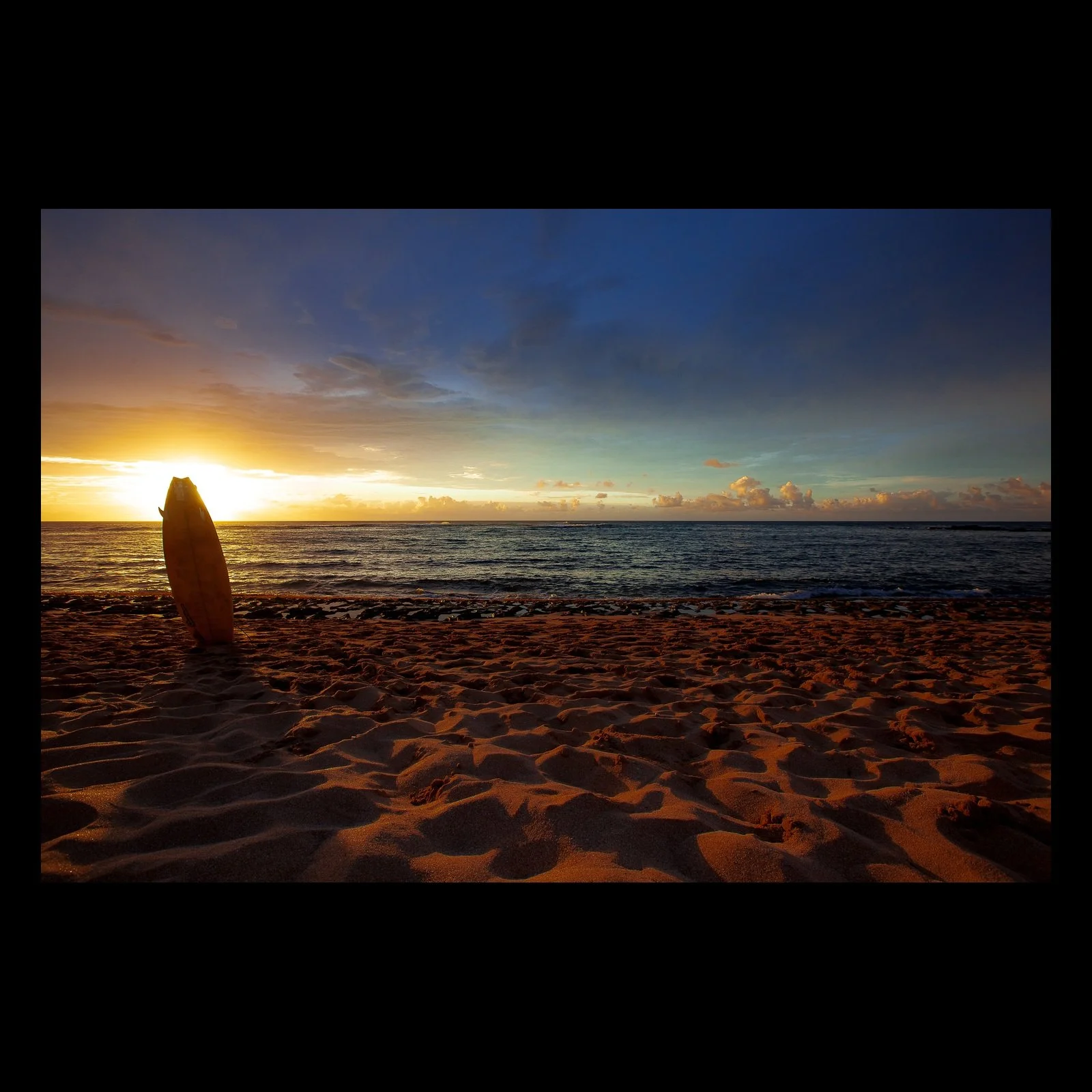 Full-frame black presentation thumbnail of Last Board Out, showing a surfboard stands upright in textured sand while the sun drops toward the ocean