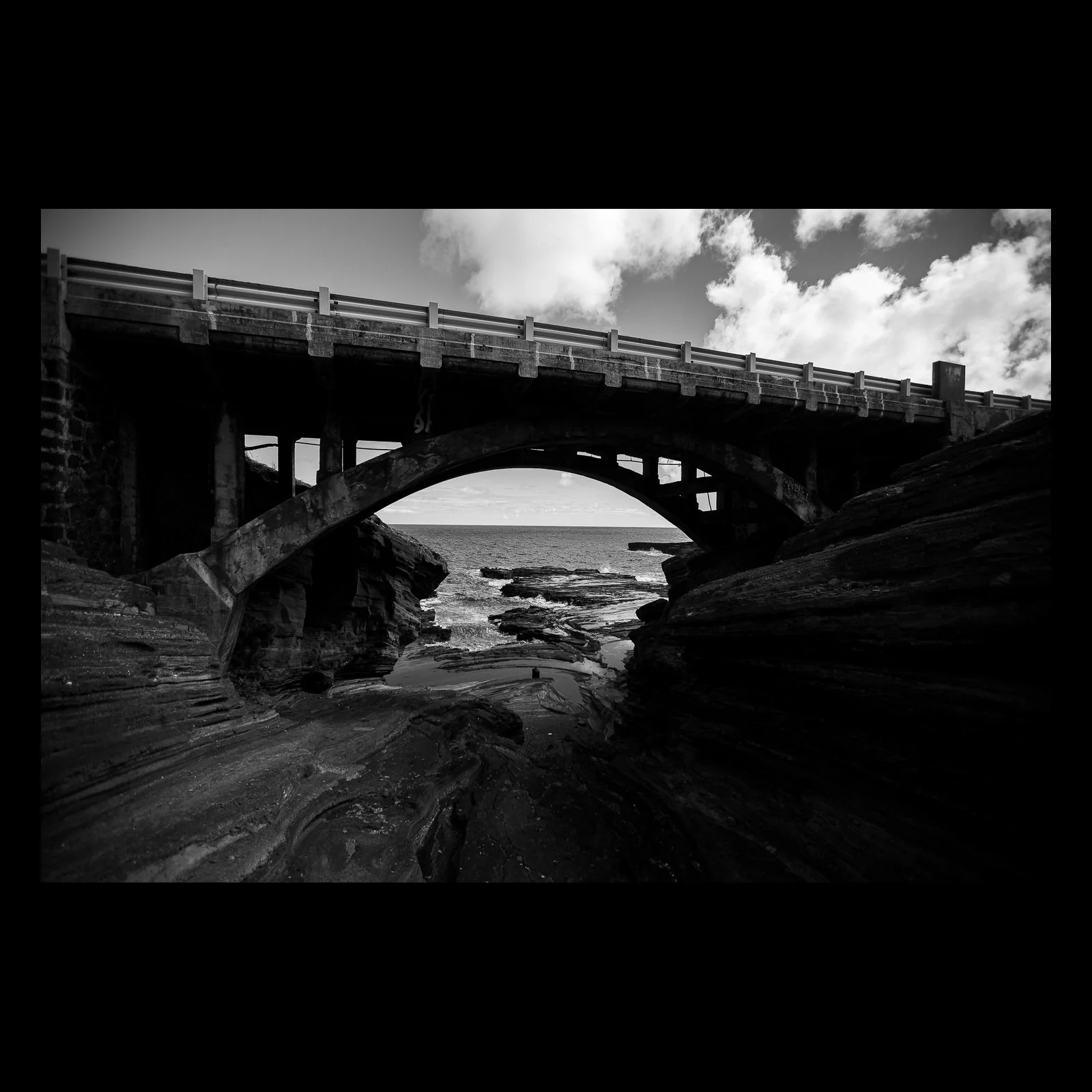 Full-frame black presentation thumbnail of Concrete Tide, showing a heavy arched bridge spans dark coastal rock and a narrow view of the sea in black and white