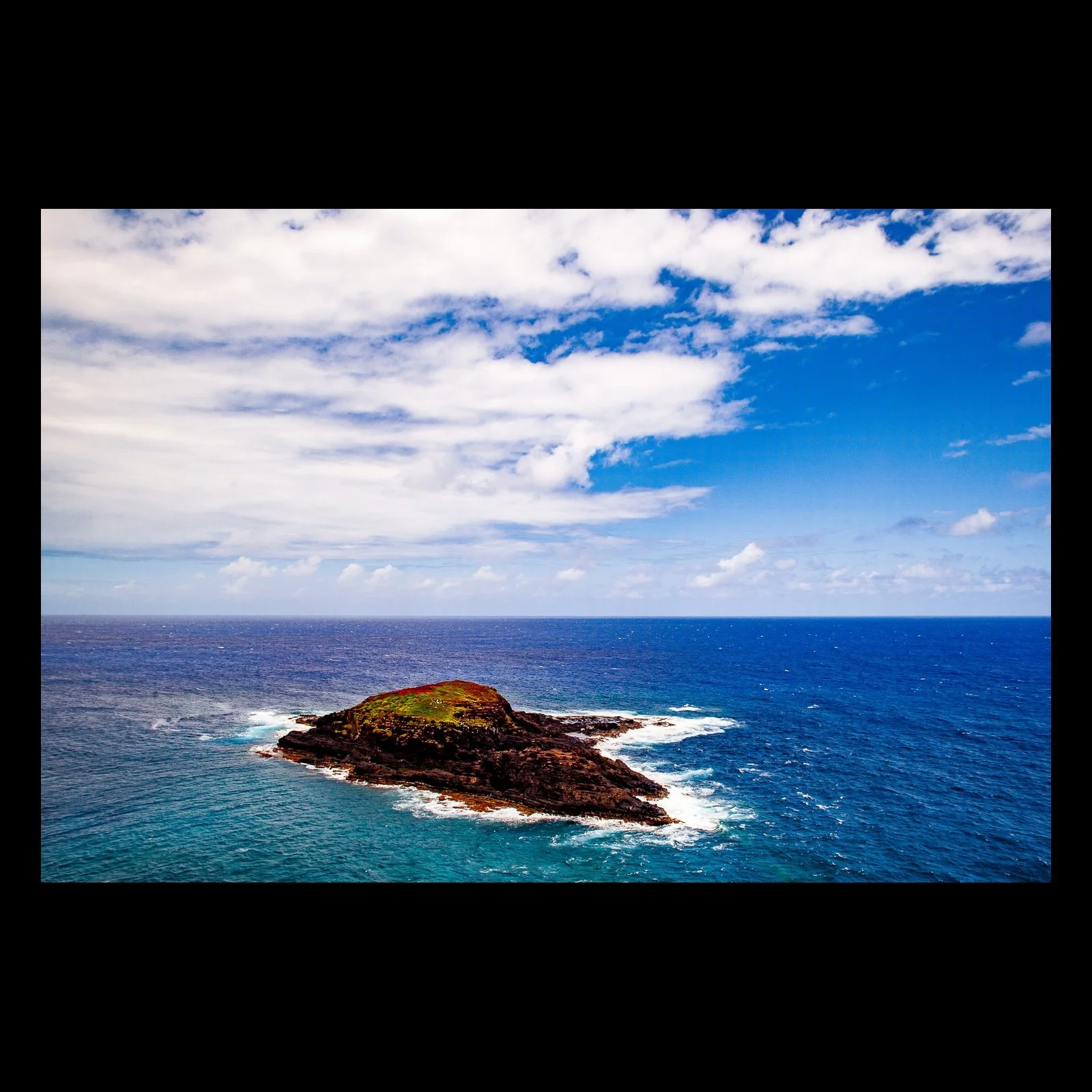 Full-frame black presentation thumbnail of Offshore Geometry, showing a low rocky island sits inside rings of white surf against a wide blue sea