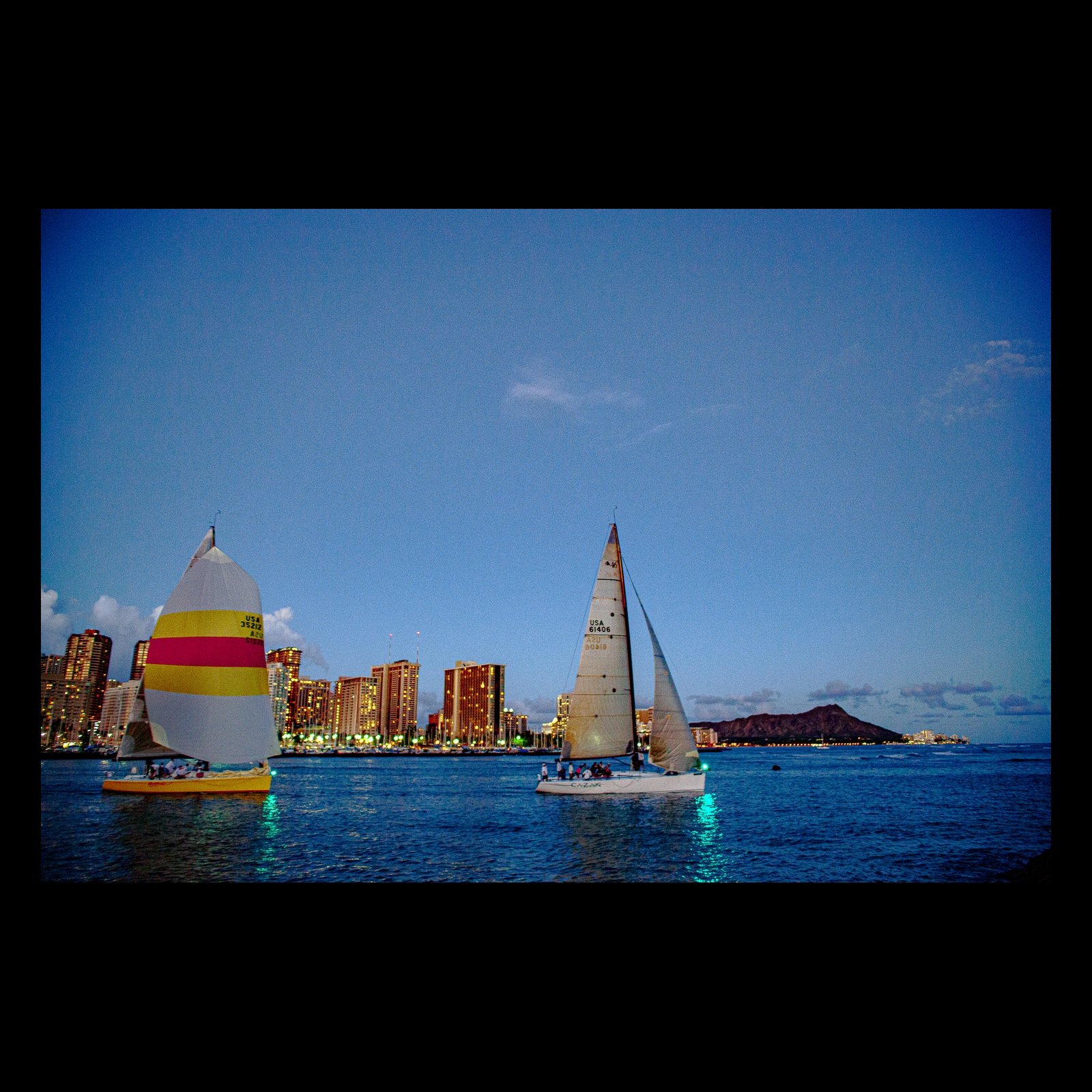 Full-frame black presentation thumbnail of Two Sails, One City, showing two sailboats cross blue water with a distant skyline and volcanic ridge behind them