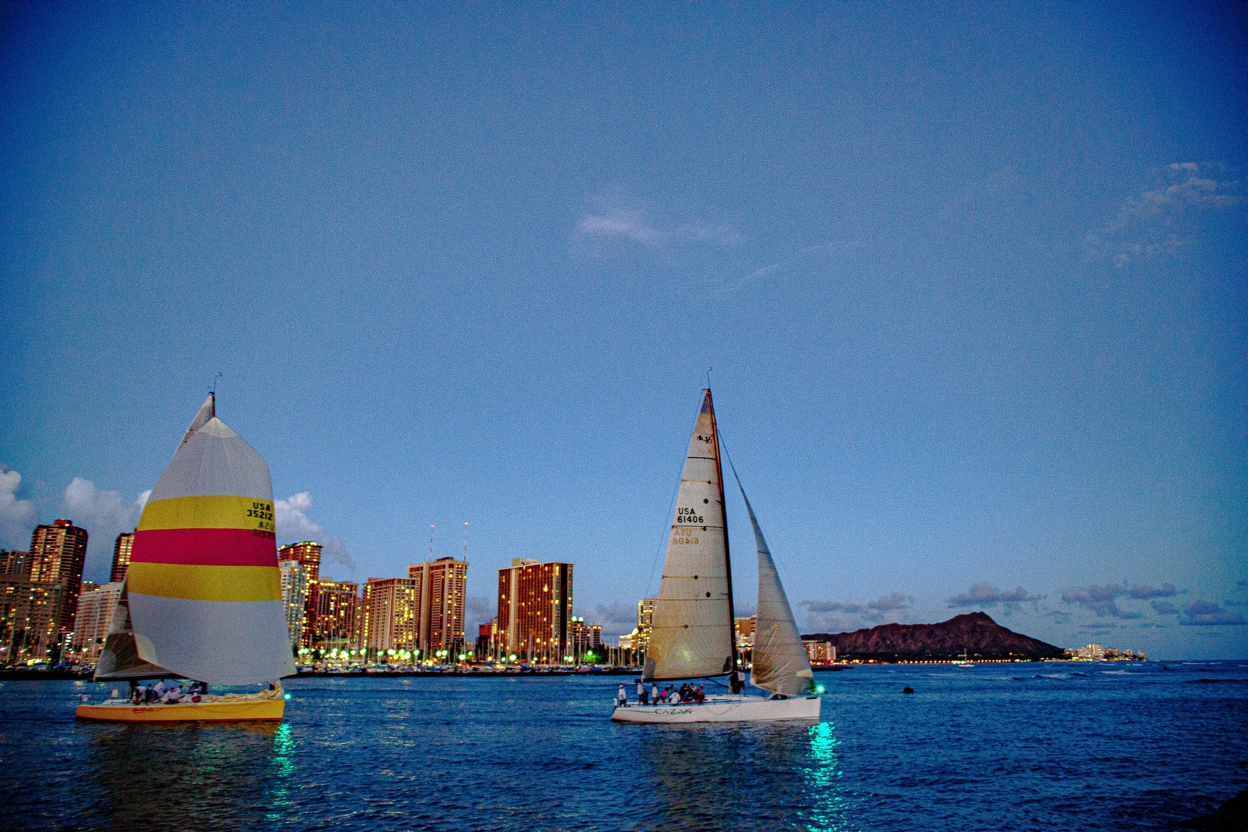 Complete uncropped artwork for Two Sails, One City, showing two sailboats cross blue water with a distant skyline and volcanic ridge behind them