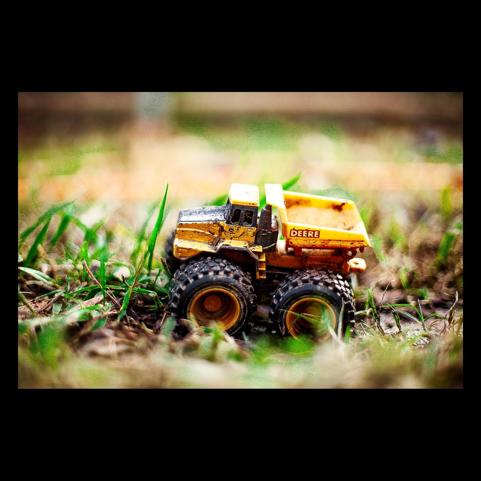 Full-frame black presentation thumbnail of Small Work, showing a small yellow toy loader sits among blades of grass at ground level