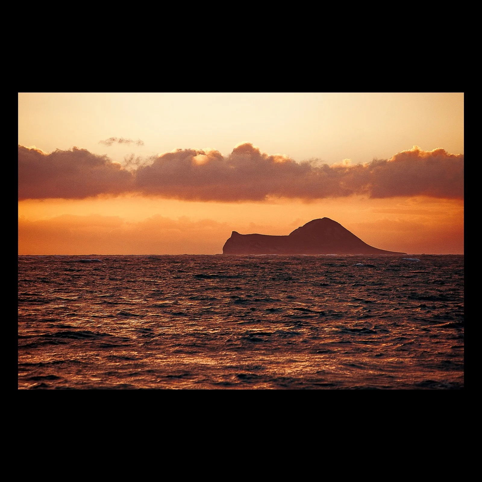 Full-frame black presentation thumbnail of Profile at Dusk, showing a dark volcanic profile sits beyond warm ocean water beneath a low band of clouds