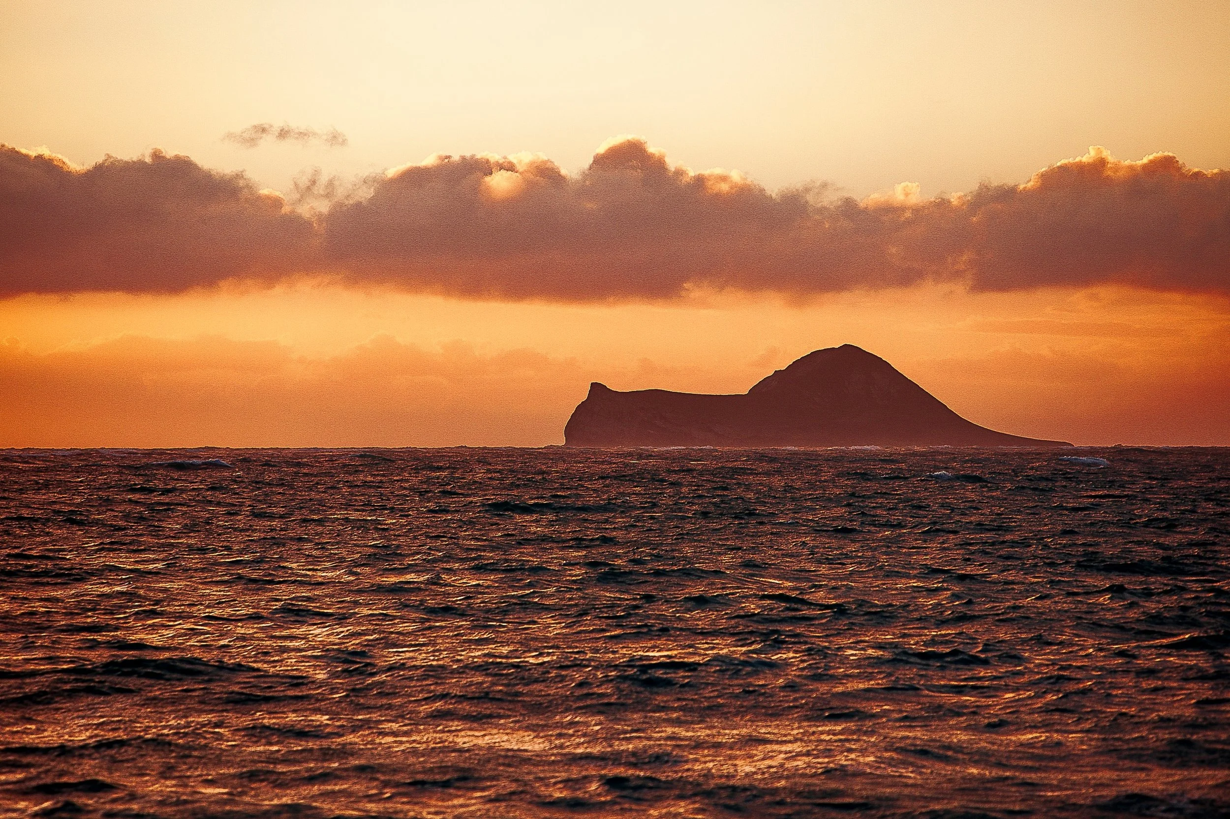 Complete uncropped artwork for Profile at Dusk, showing a dark volcanic profile sits beyond warm ocean water beneath a low band of clouds