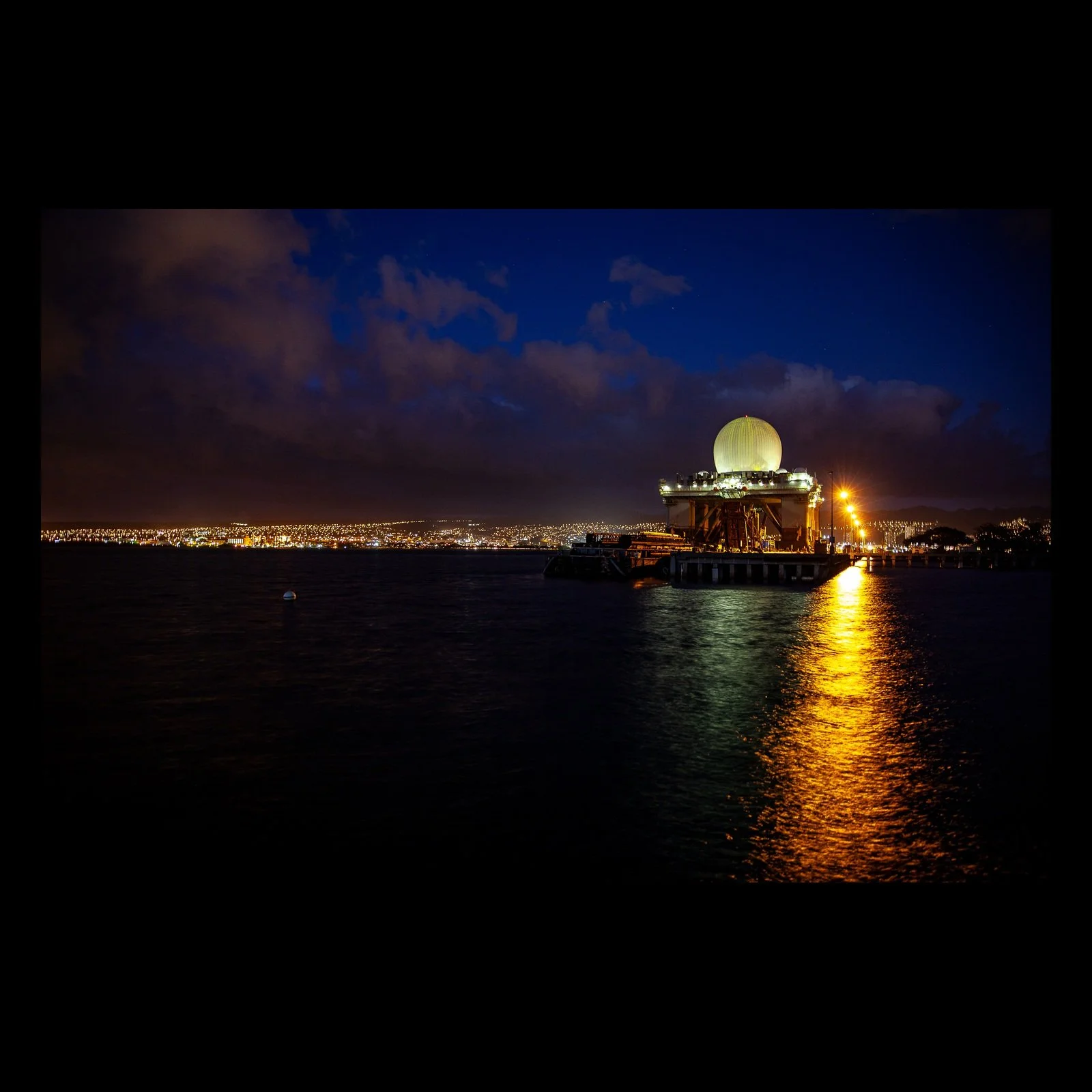 Full-frame black presentation thumbnail of Night Beacon, showing a luminous waterfront structure casts a long gold reflection across dark water at night