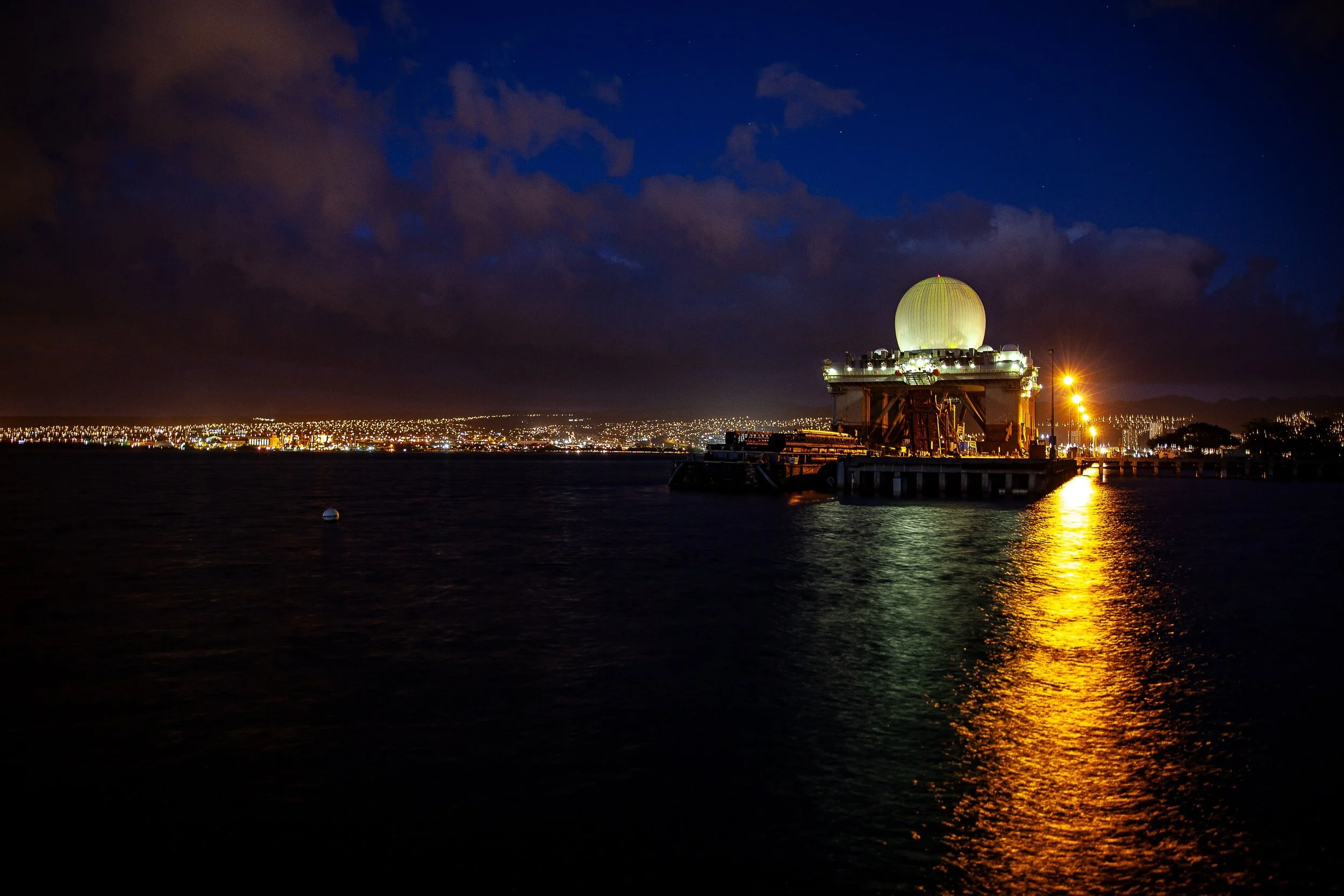 Complete uncropped artwork for Night Beacon, showing a luminous waterfront structure casts a long gold reflection across dark water at night