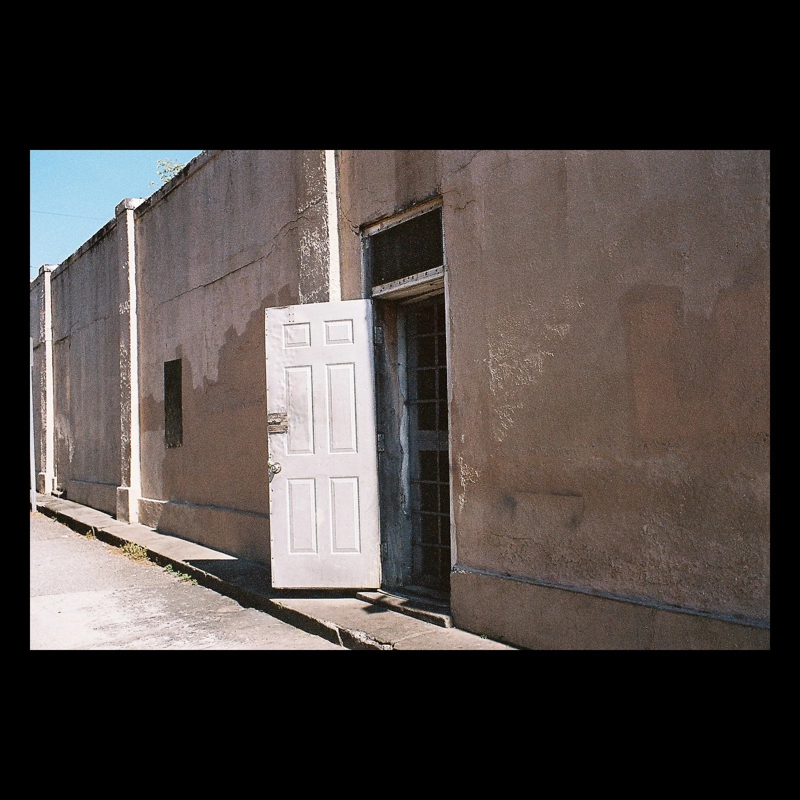 Full-frame black presentation thumbnail of The Open Door, showing a white door stands open in a sunlit wall marked by age and peeling surfaces