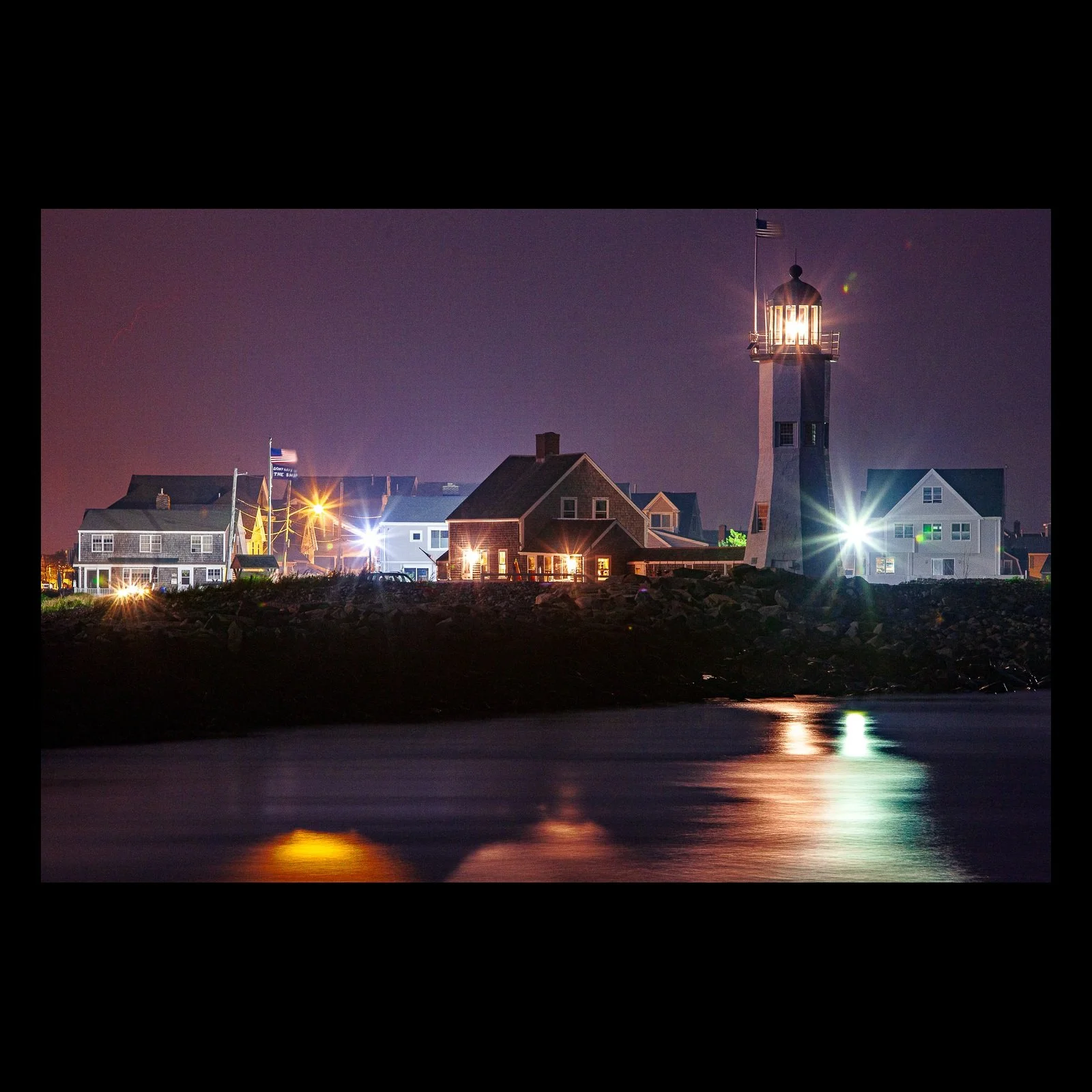 Full-frame black presentation thumbnail of Harbor Lantern, showing a lighthouse and clustered coastal buildings shine across dark water at night