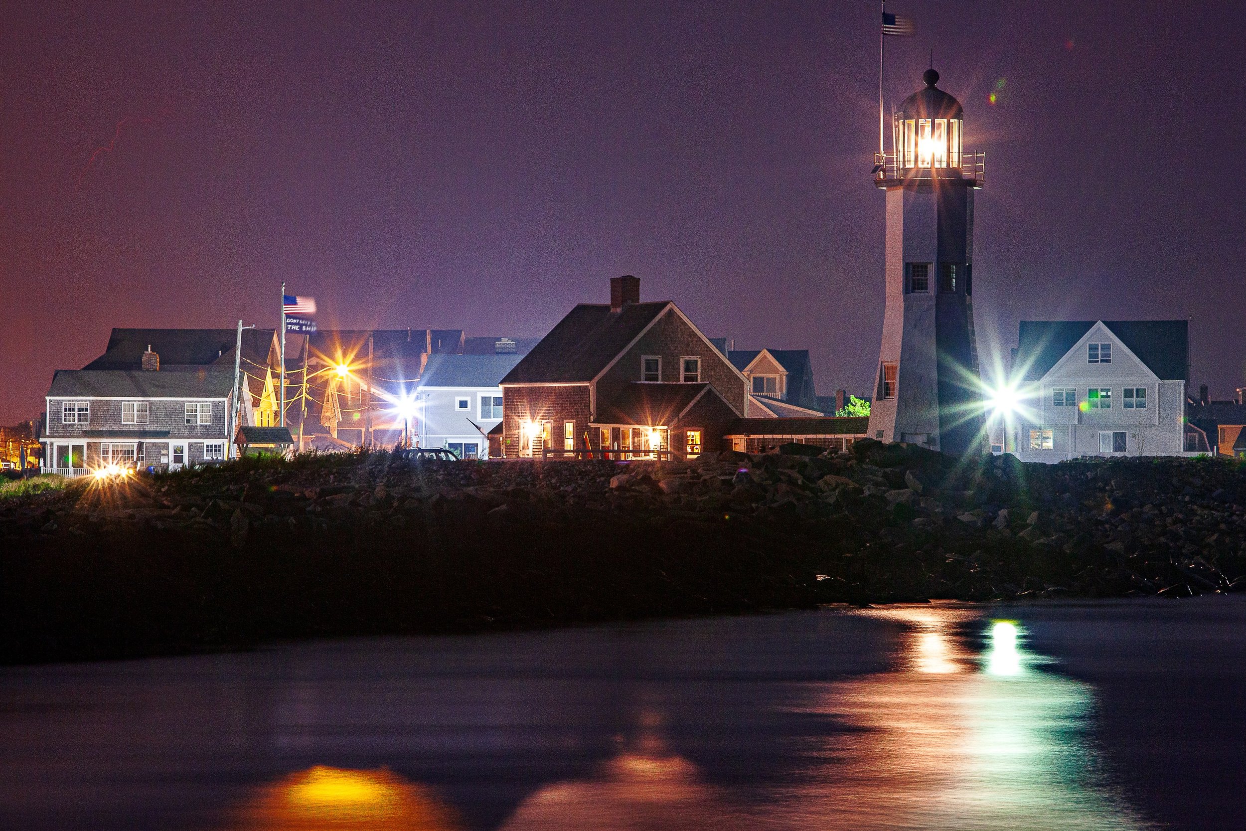 Complete uncropped artwork for Harbor Lantern, showing a lighthouse and clustered coastal buildings shine across dark water at night