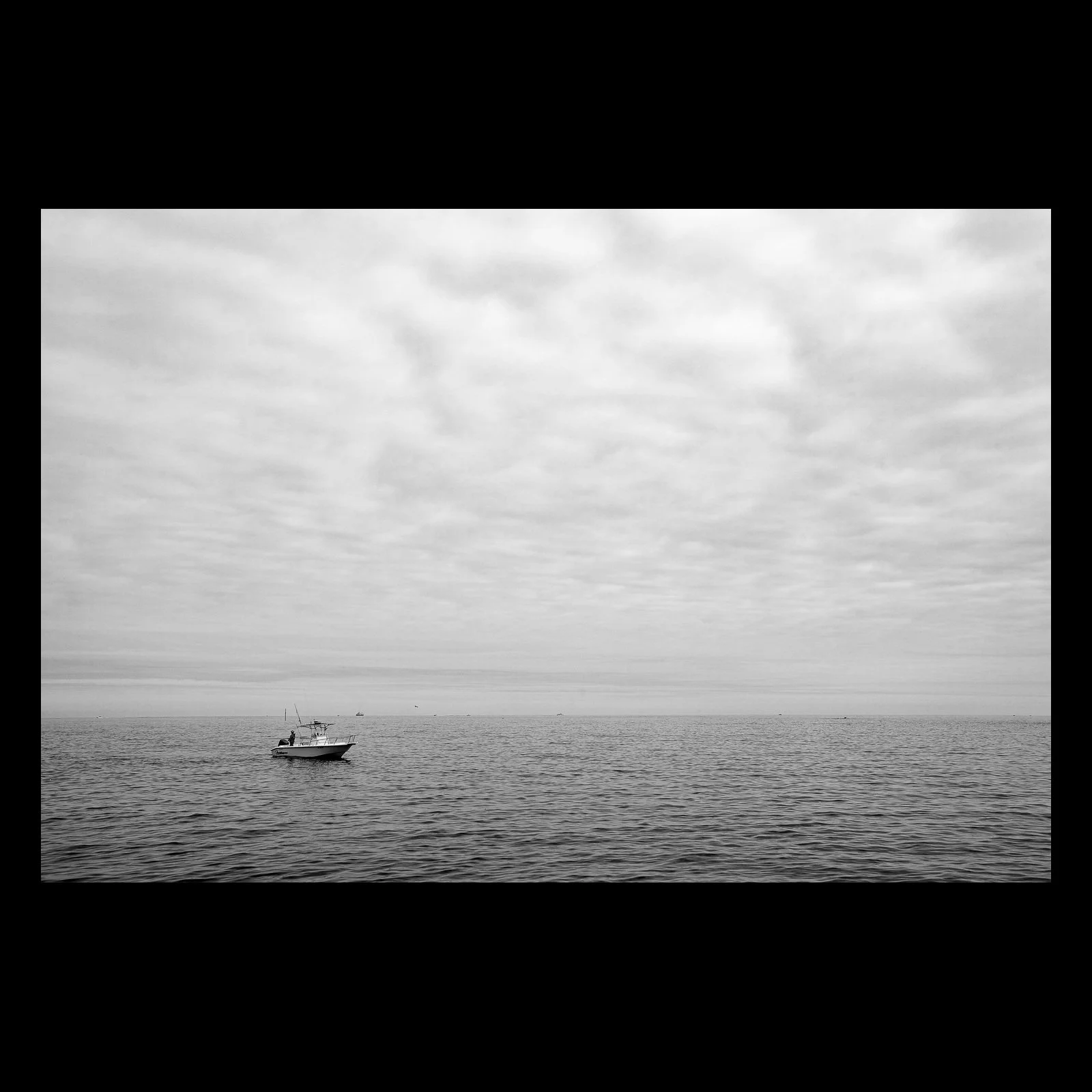 Full-frame black presentation thumbnail of A Boat in the Blank, showing a single small boat rests beneath an expansive gray sky and faint horizon in black and white