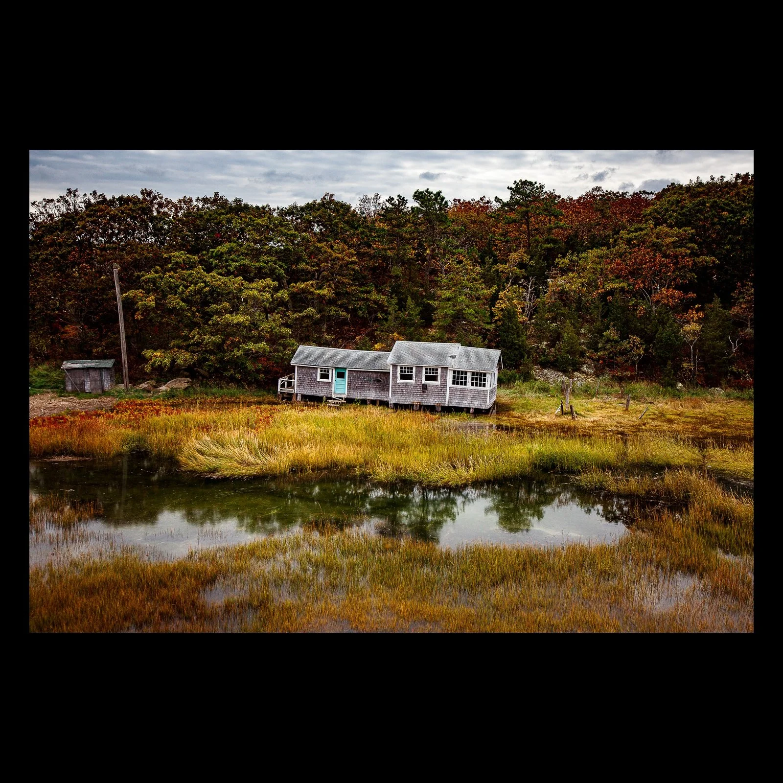 Full-frame black presentation thumbnail of Salt Marsh Quiet, showing a weathered cottage reflects in still marsh water beneath autumn-colored trees