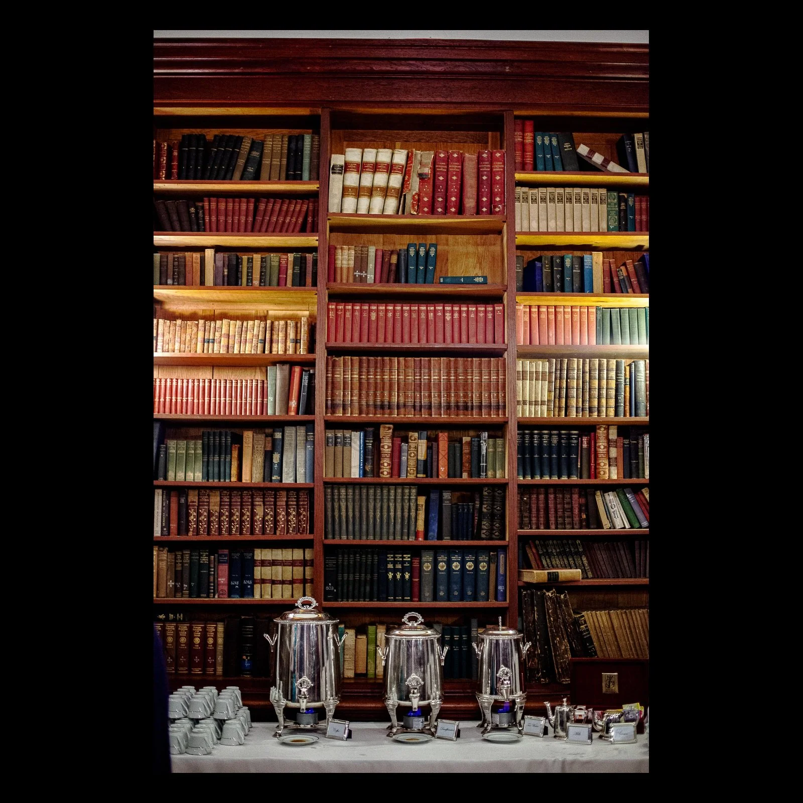 Full-frame black presentation thumbnail of The Quiet Library, showing tall shelves of richly colored books rise behind a formal table in a vertical interior