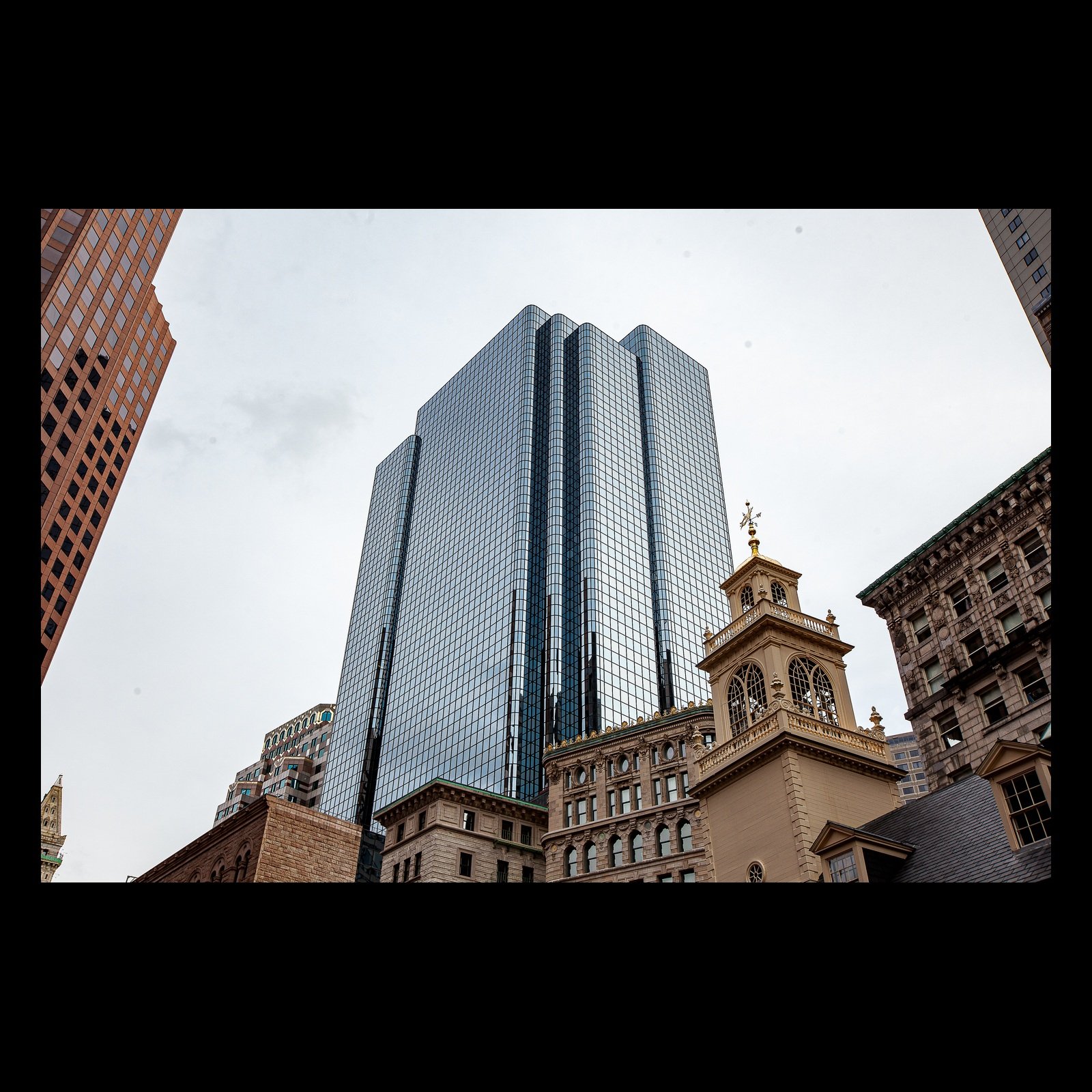 Full-frame black presentation thumbnail of Old and New, showing a reflective modern tower rises between older stone and brick buildings in a low-angle city view