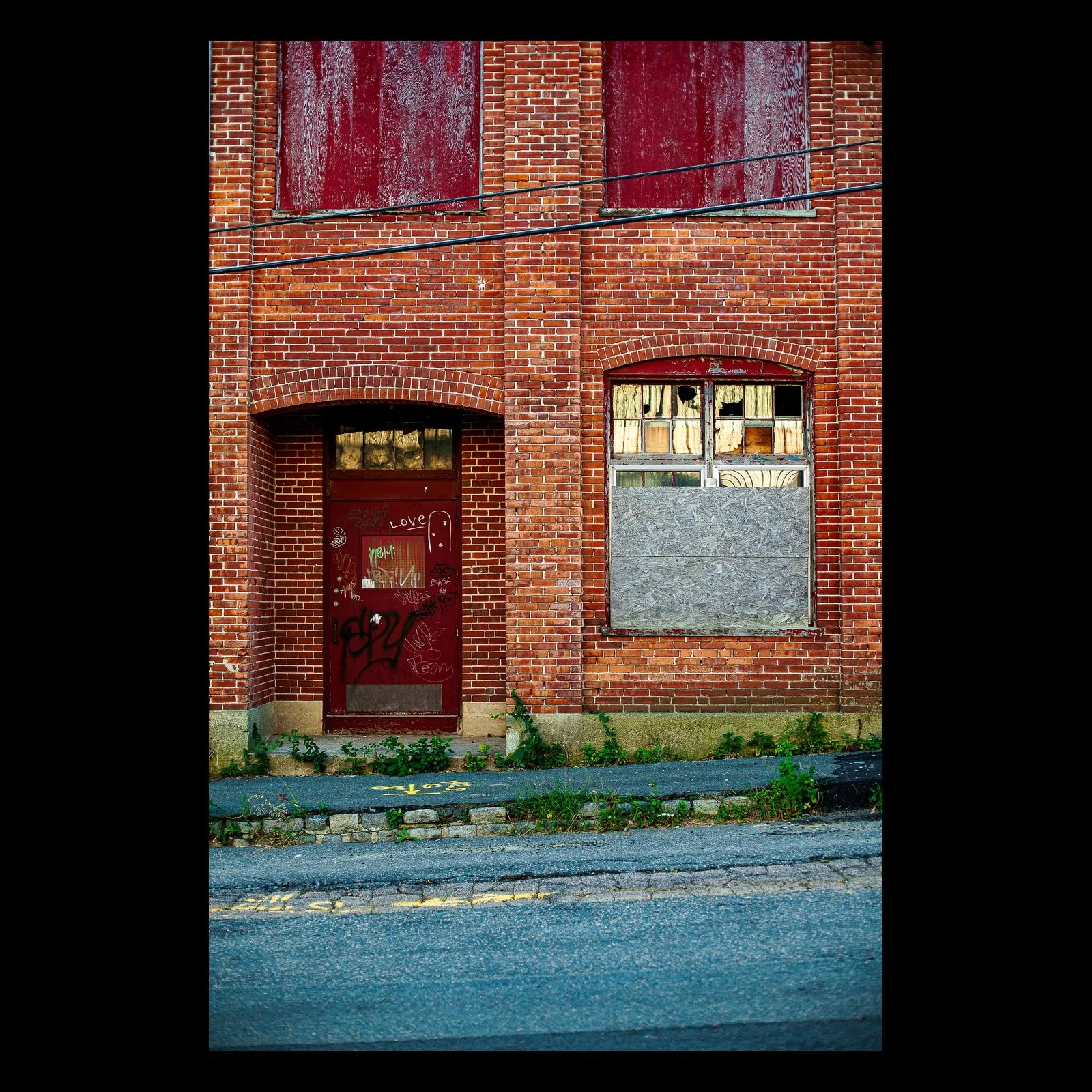 Full-frame black presentation thumbnail of Tainted Love, showing a red door and boarded window punctuate an aging brick facade
