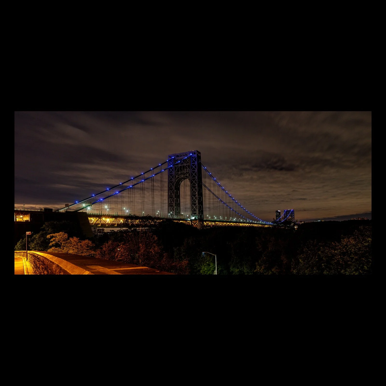Full-frame black presentation thumbnail of Blue Suspension, showing a long suspension bridge glows blue above sweeping highway lights beneath a clouded night sky