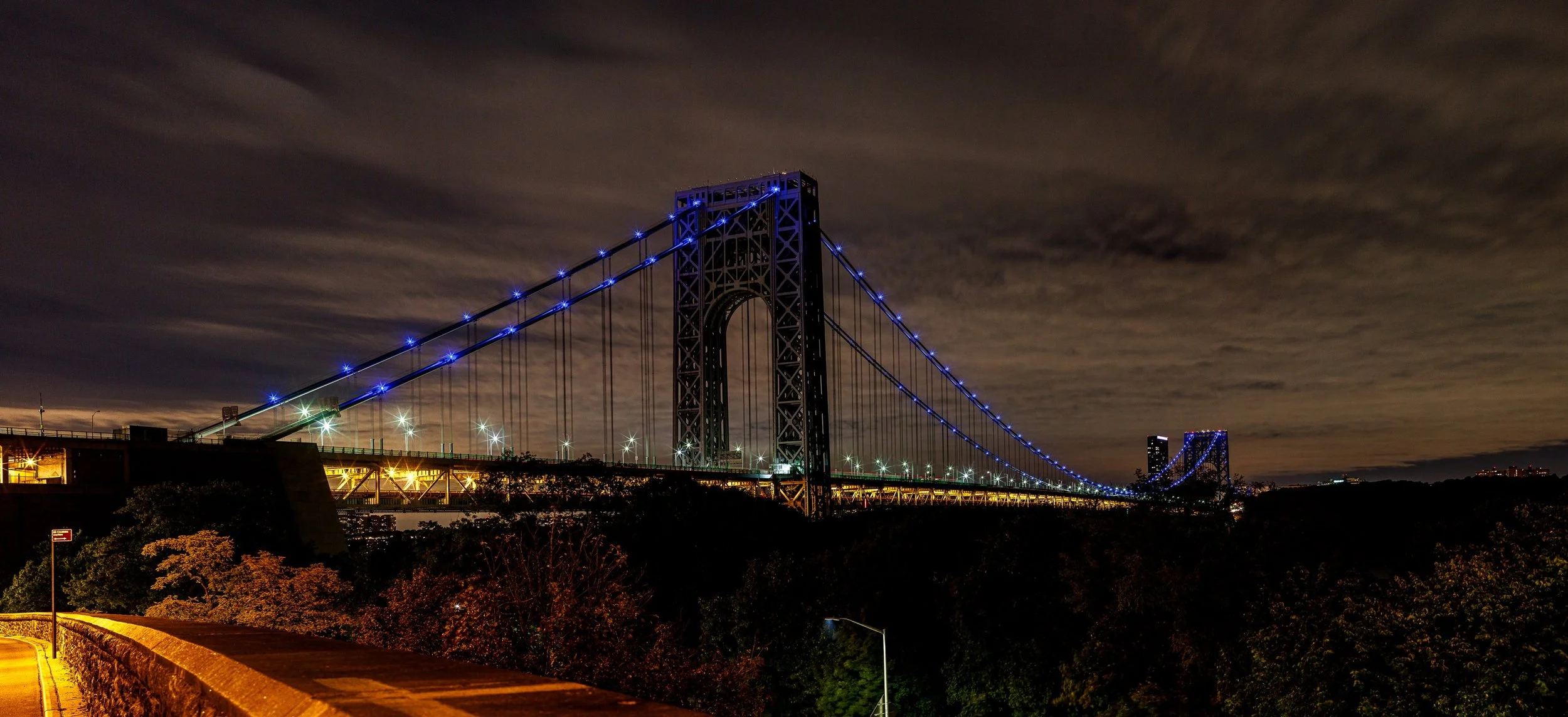 Complete uncropped artwork for Blue Suspension, showing a long suspension bridge glows blue above sweeping highway lights beneath a clouded night sky