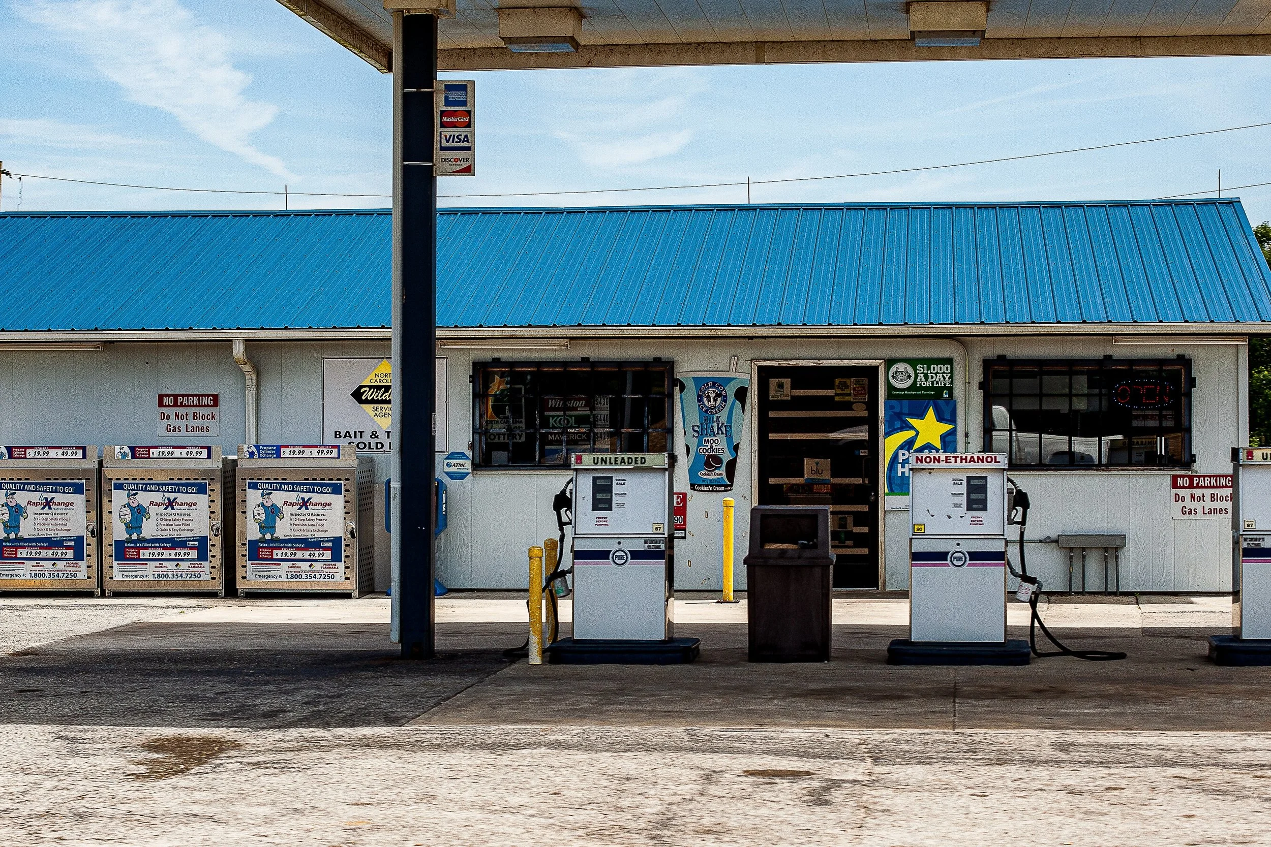 Detail crop from Last Fuel, emphasizing pumps, signs, stains, and parked cars reveal the building's working life