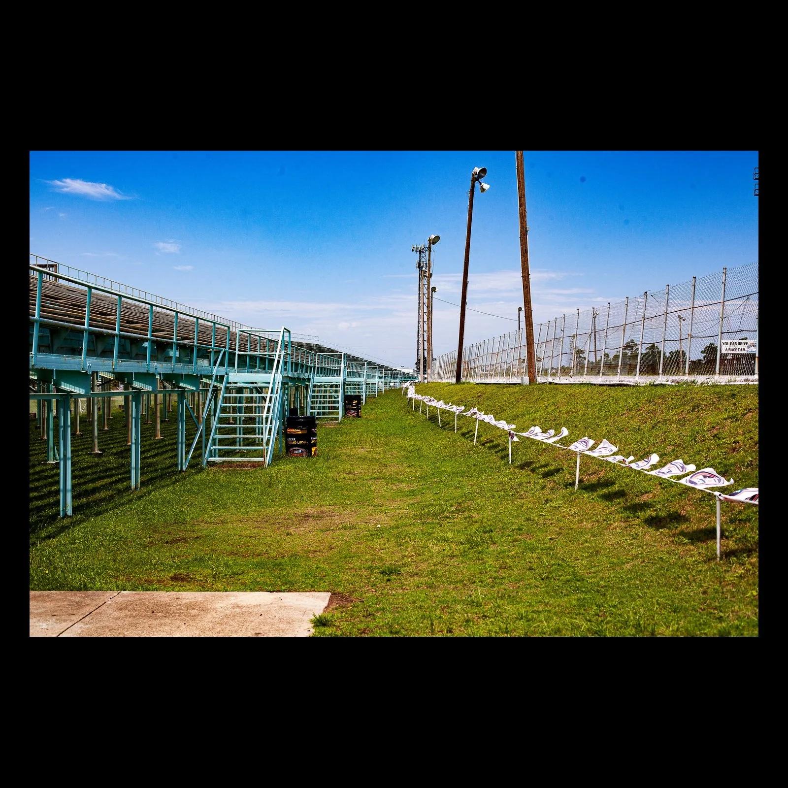 Full-frame black presentation thumbnail of Grandstand After Summer, showing empty bleachers and a line of folded seats stretch beside a green field under clear sky