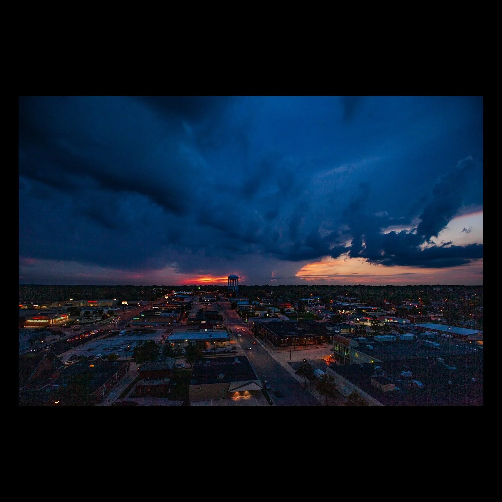 Full-frame black presentation thumbnail of Storm Over Town, showing a dark blue storm shelf advances above a town while a thin red sunset remains at the horizon