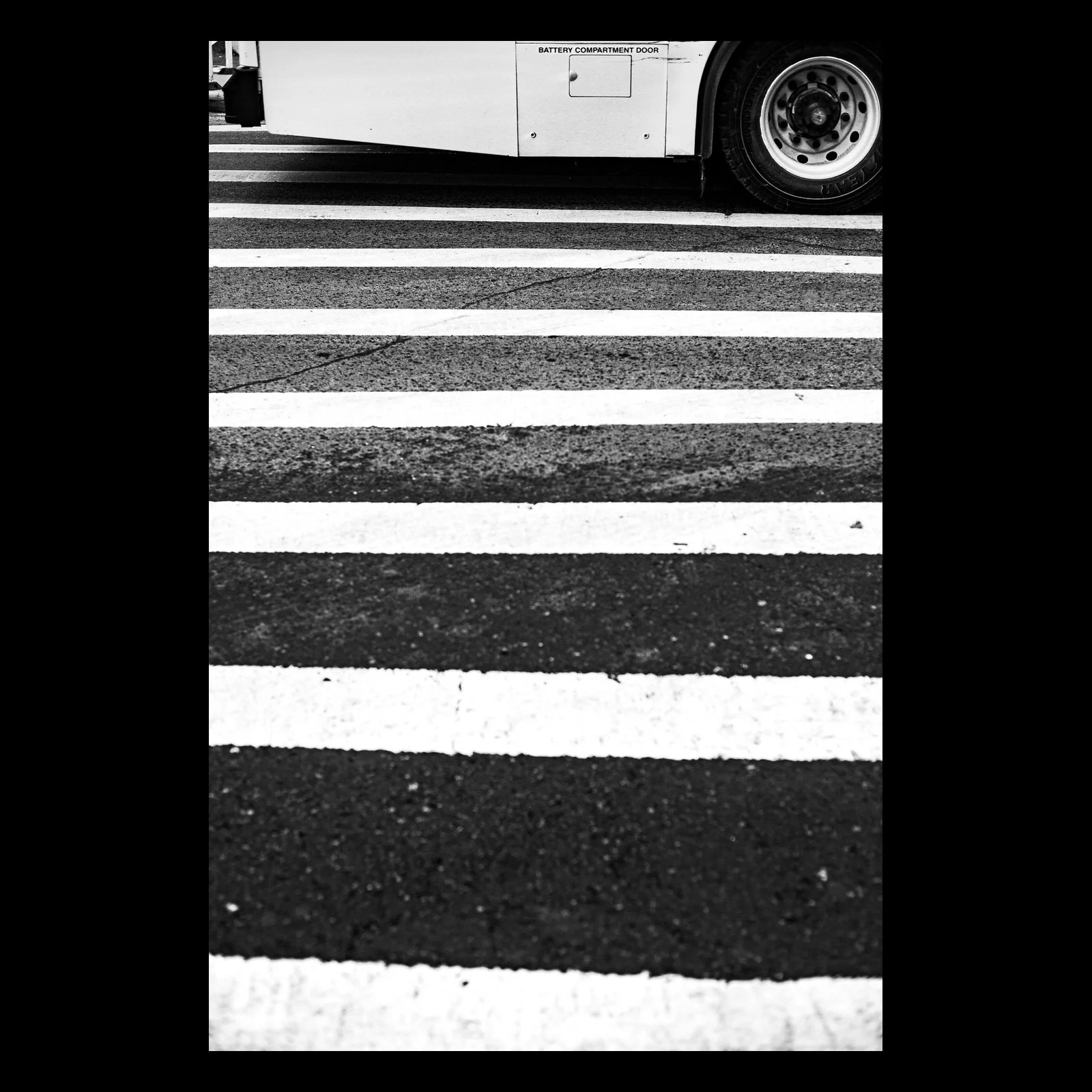 Full-frame black presentation thumbnail of Crossing Lines, showing a bus tire meets bold white crosswalk stripes in a narrow vertical street view