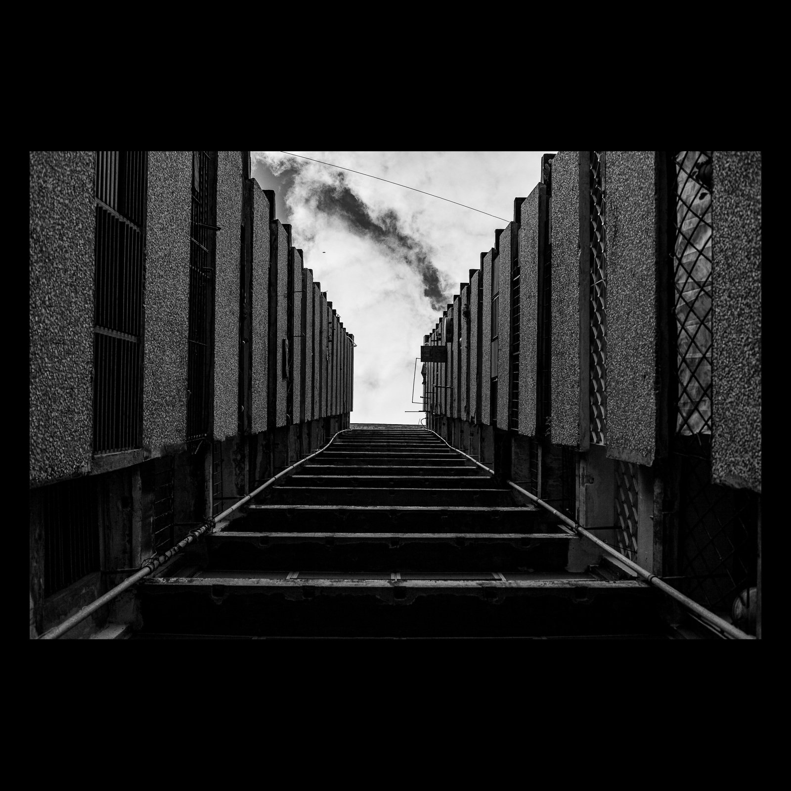 Full-frame black presentation thumbnail of Industrial Ascent, showing a steep service stair climbs between repeating industrial walls toward a plume of steam