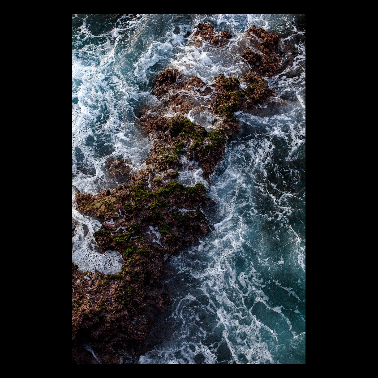 Full-frame black presentation thumbnail of Sea Against Stone, showing turquoise water and white foam churn around dark coastal rock in an overhead vertical view