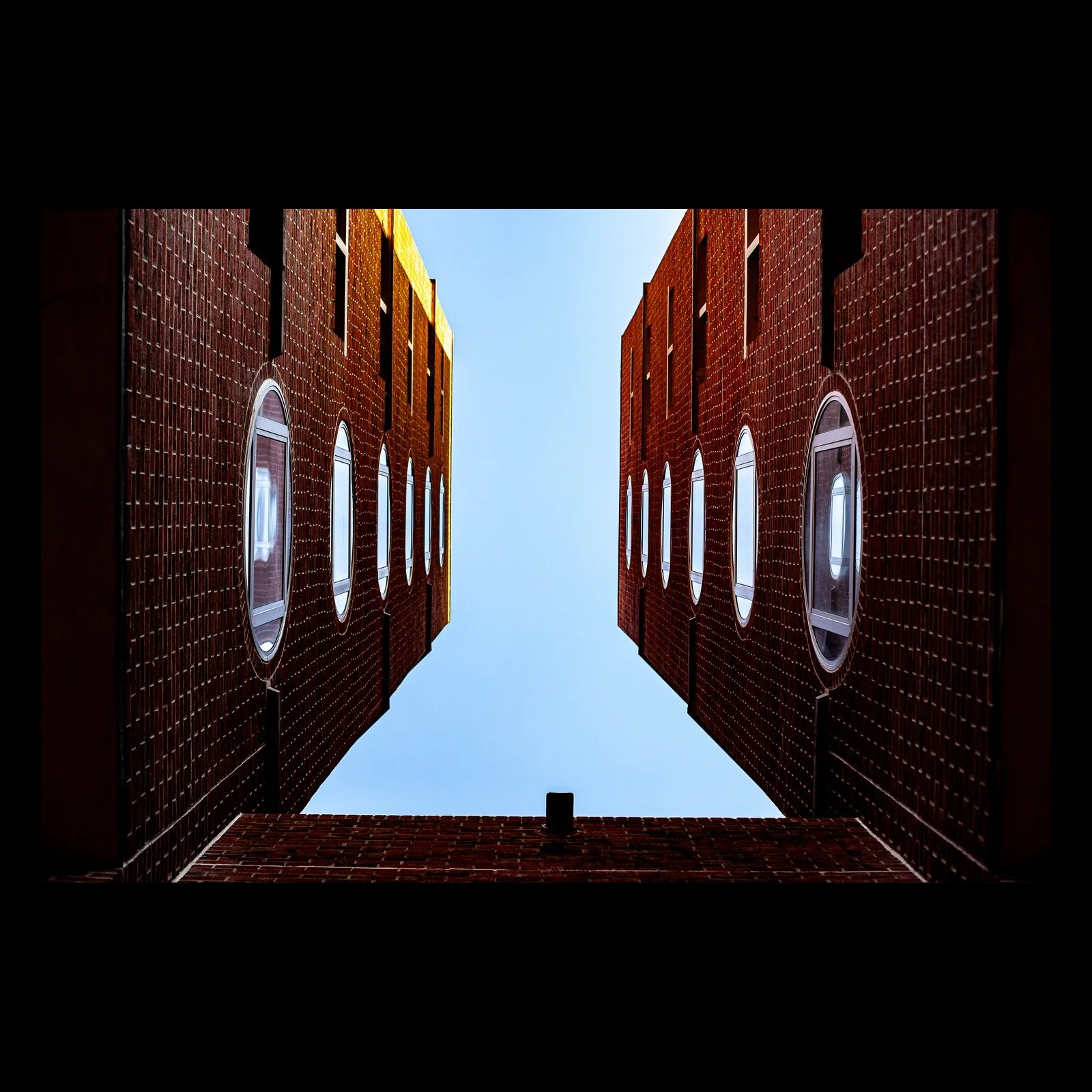 Full-frame black presentation thumbnail of Sky Courtyard, showing three red-brick walls and oval windows frame a clean opening of blue sky