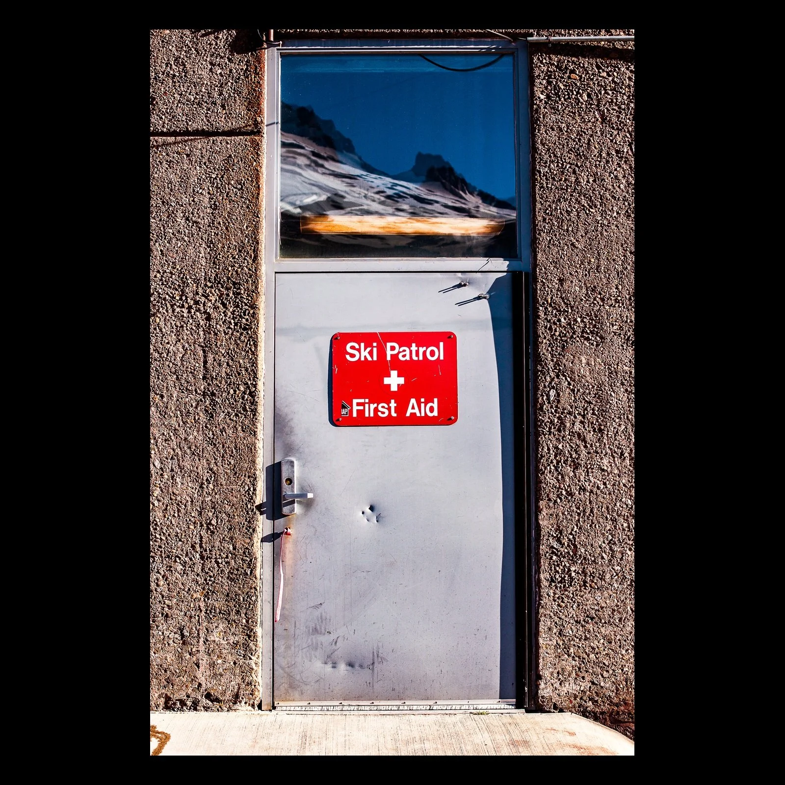 Full-frame black presentation thumbnail of First Aid, showing a utilitarian door marked for ski patrol and first aid stands beneath a small mountain image
