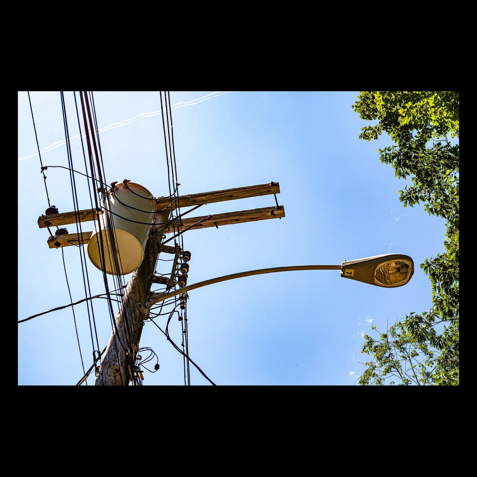 Full-frame black presentation thumbnail of Utility Sky, showing power lines, transformers, and a streetlight cross a bright blue sky beside green leaves