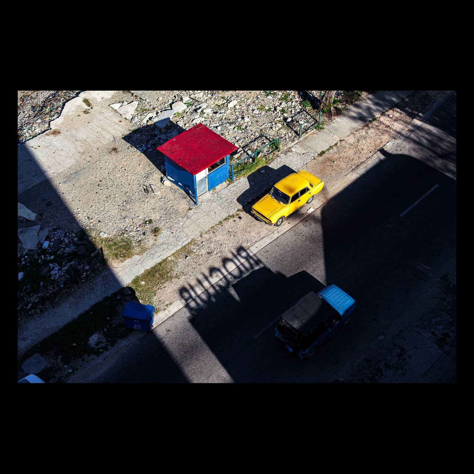 Full-frame black presentation thumbnail of Primary Geometry, showing a yellow car and blue-roofed structure sit inside strong blocks of shadow viewed from above