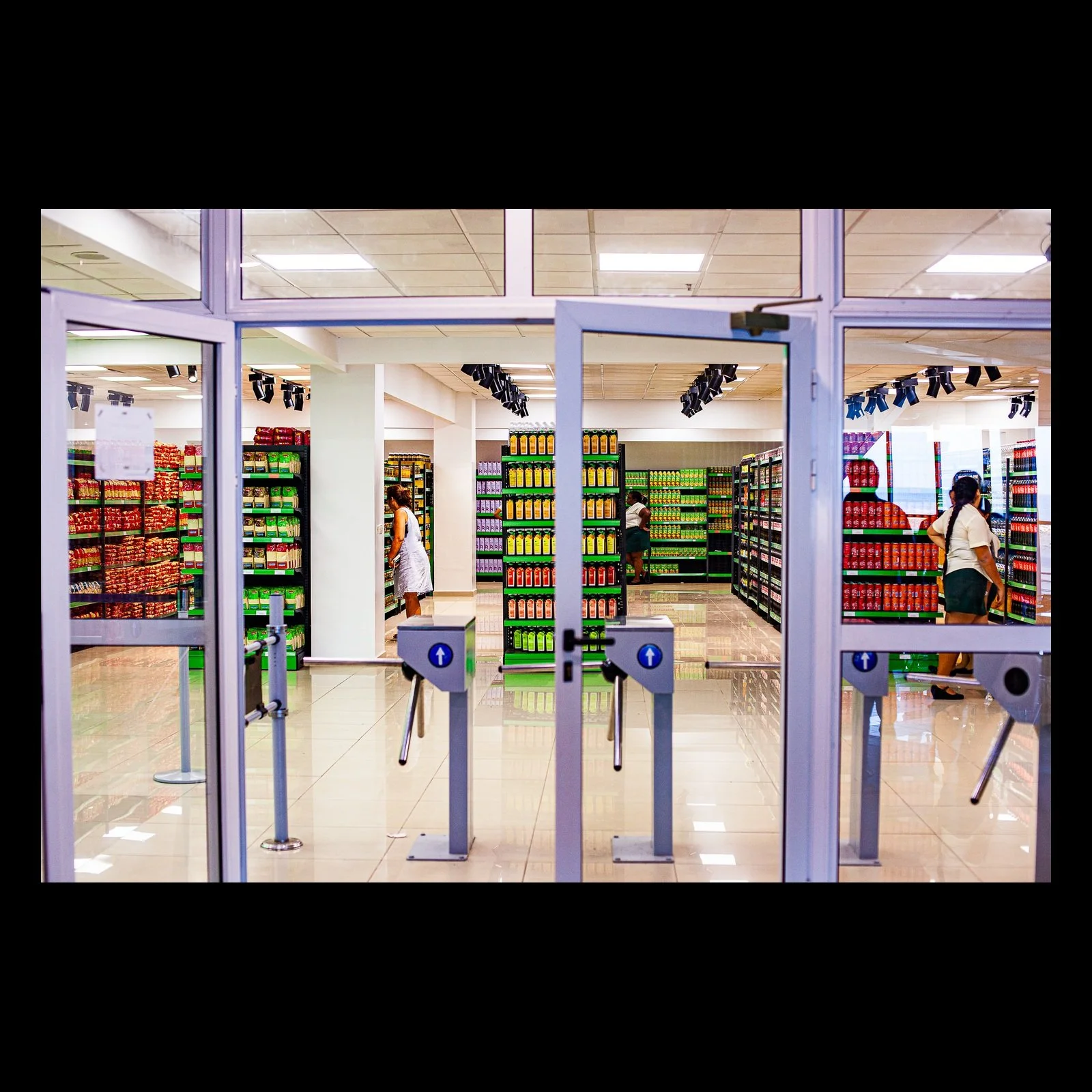 Full-frame black presentation thumbnail of The Threshold, showing glass entry gates frame a brightly ordered shop interior filled with colorful rows