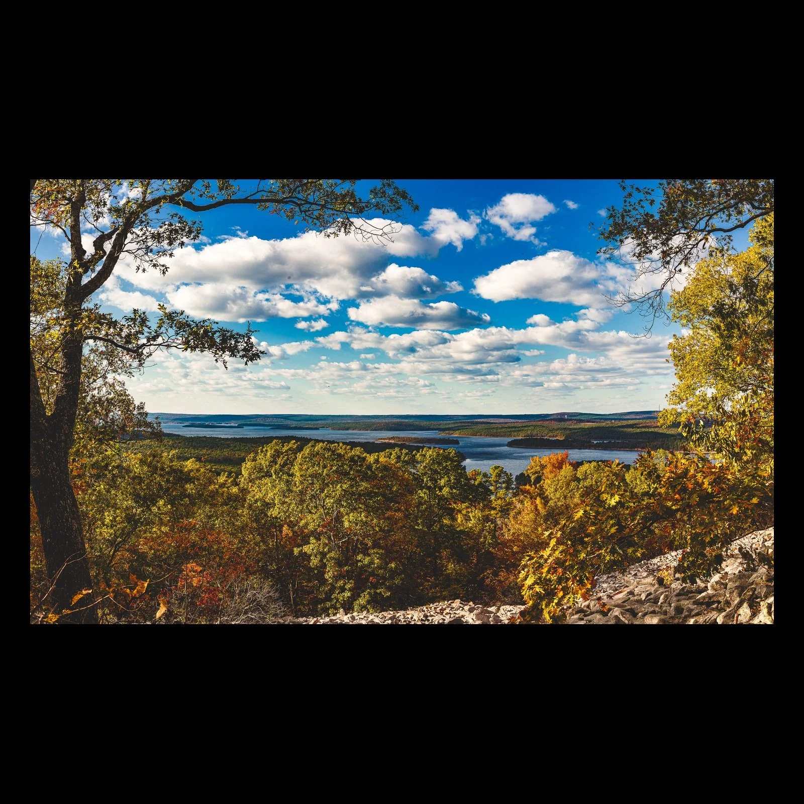 Full-frame black presentation thumbnail of Autumn Lookout, showing golden trees frame a broad lake and distant rolling terrain under blue sky and white clouds