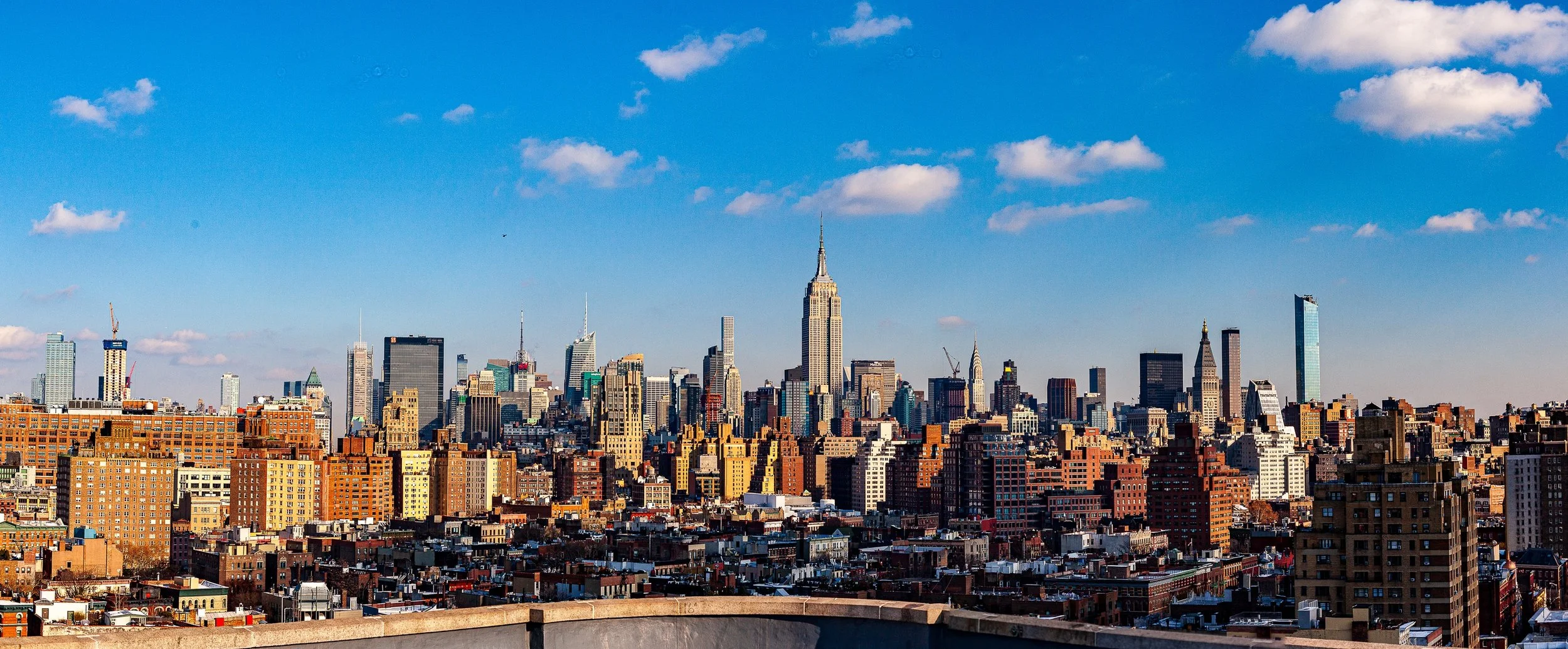 Complete uncropped artwork for Empire Distance, showing a dense skyline rises beyond brick neighborhoods under a clear blue sky