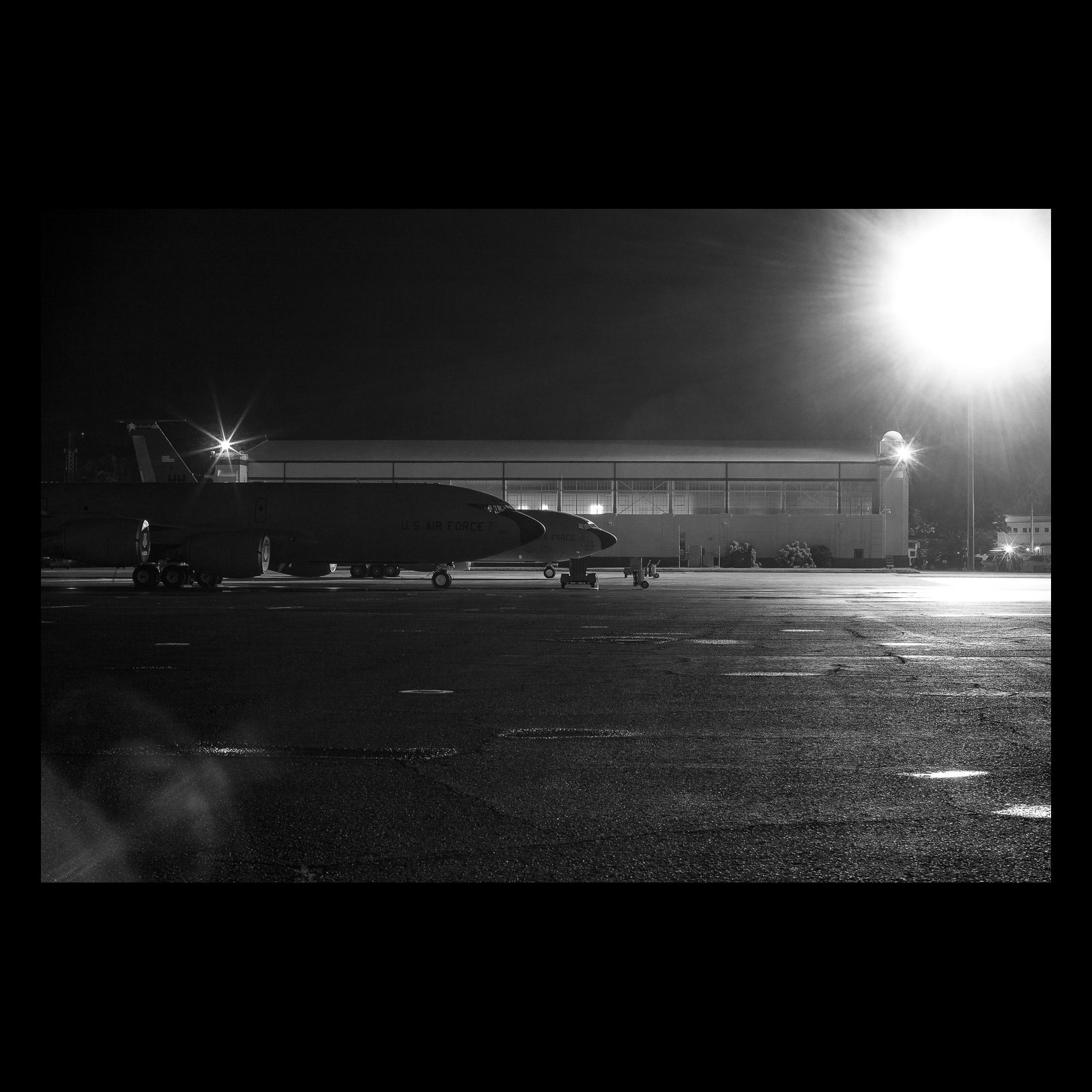 Full-frame black presentation thumbnail of Flightline Noir, showing a military aircraft rests beneath intense ramp lights in a grainy black-and-white night scene