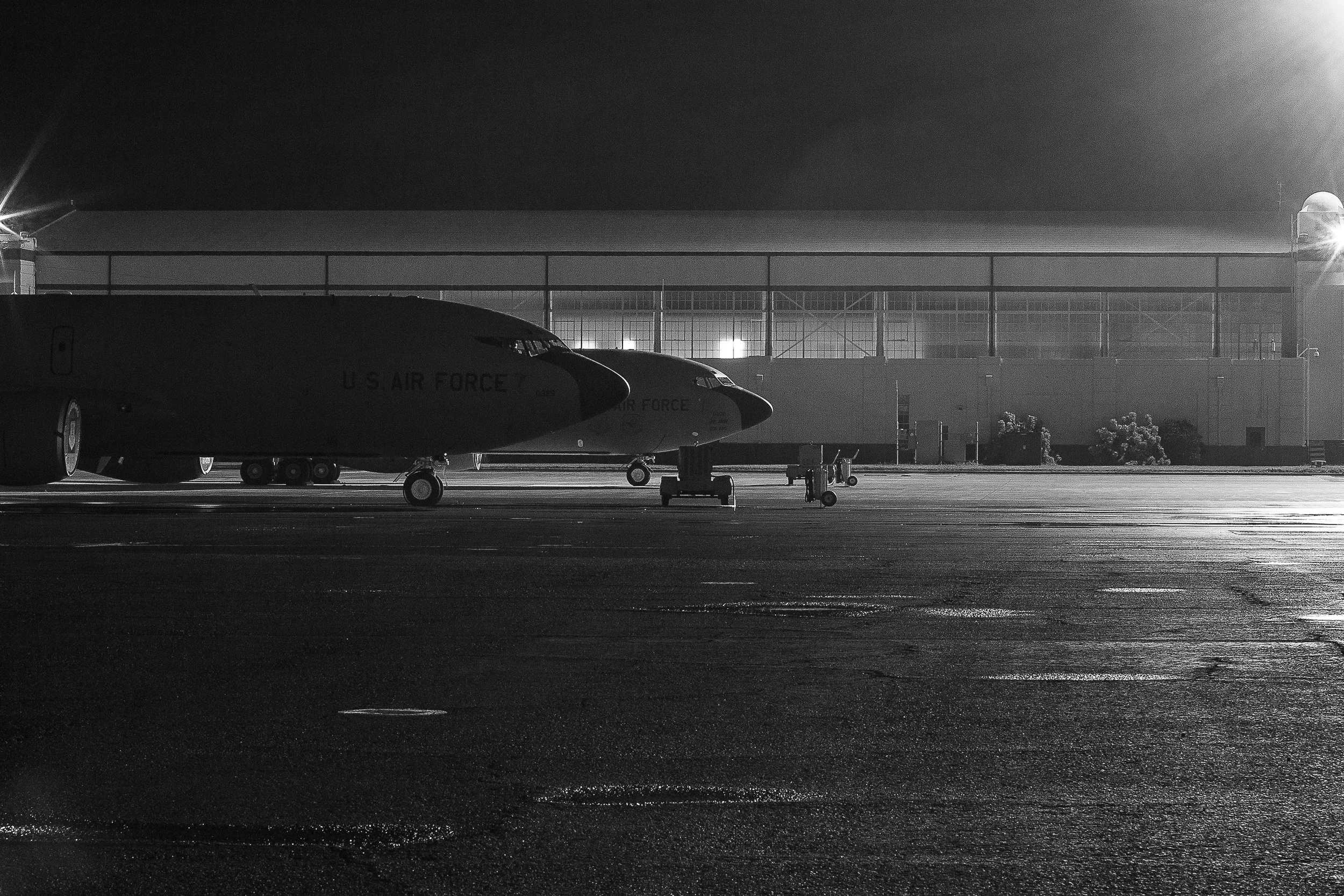 Detail crop from Flightline Noir, emphasizing wet pavement, distant structures, and faint personnel give depth to the flight line